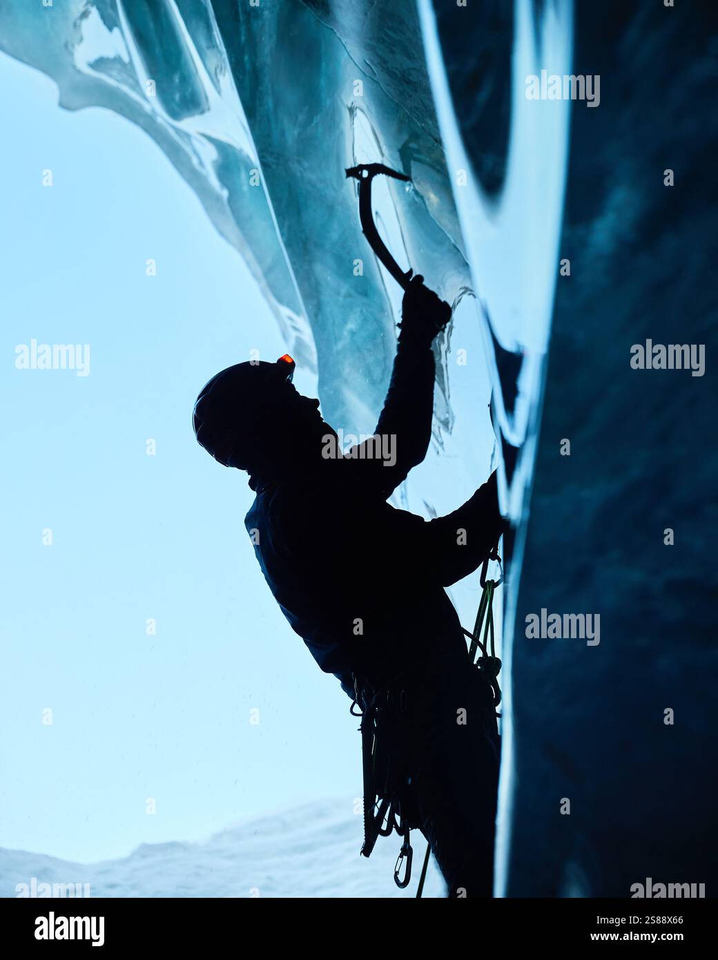 Ein Mann in schwarzer Silhouette klettert auf eine Eishöhle im Inneren eines großen Gletschers mit Eisaxt und Steigeisen gegen blaue Ski in den Winterbergen. Sport-Mo Stockfoto