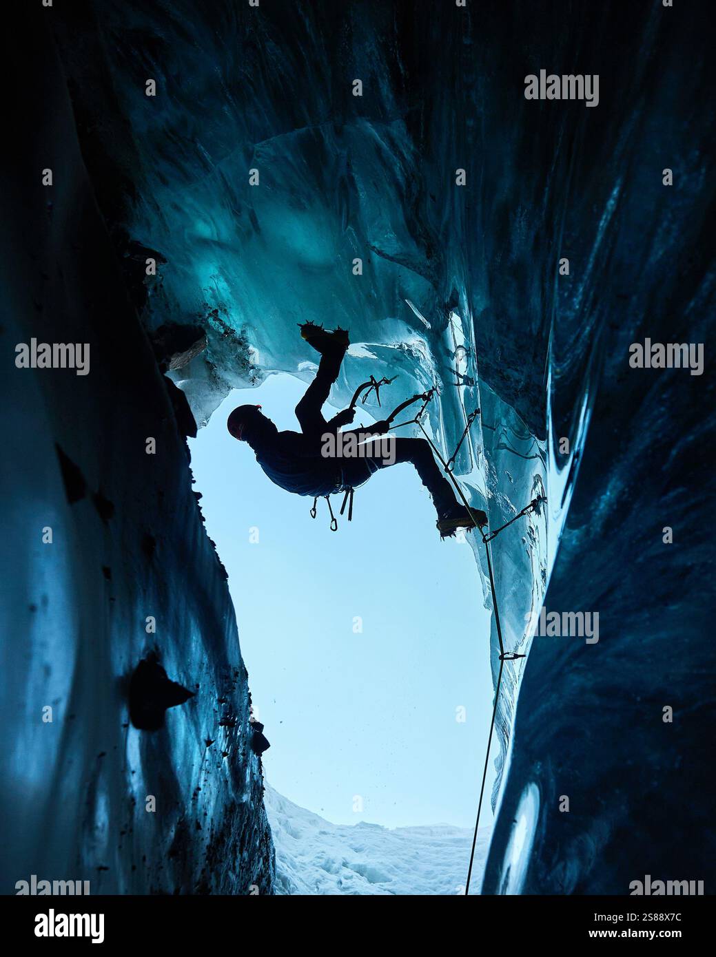 Ein Mann in schwarzer Silhouette klettert auf eine Eishöhle im Inneren eines großen Gletschers mit Eisaxt und Steigeisen gegen blaue Ski in den Winterbergen. Sport-Mo Stockfoto