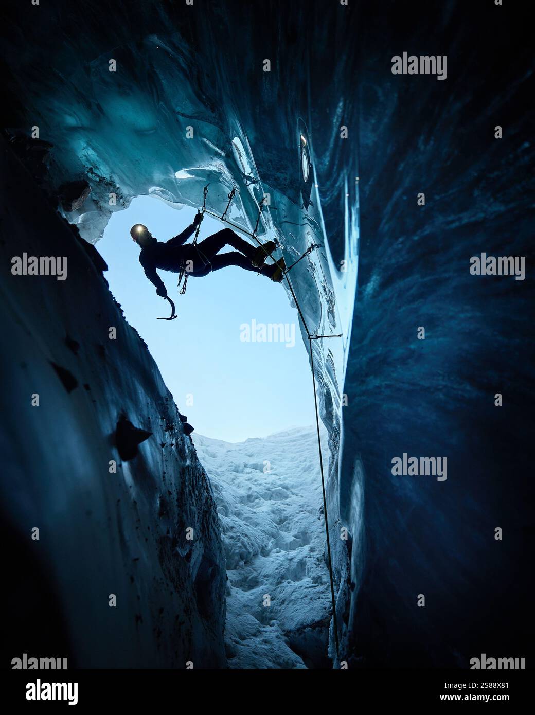 Ein Mann in schwarzer Silhouette klettert auf eine Eishöhle im Inneren eines großen Gletschers mit Eisaxt und Steigeisen gegen blaue Ski in den Winterbergen. Sport-Mo Stockfoto