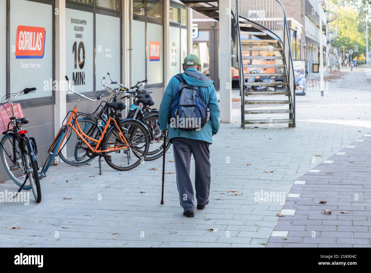 Ein alter Mann mit Rucksack und Stock läuft mühsam auf dem Bürgersteig einer verlassenen Einkaufsstraße in einem Vorort von Amsterdam. Stockfoto