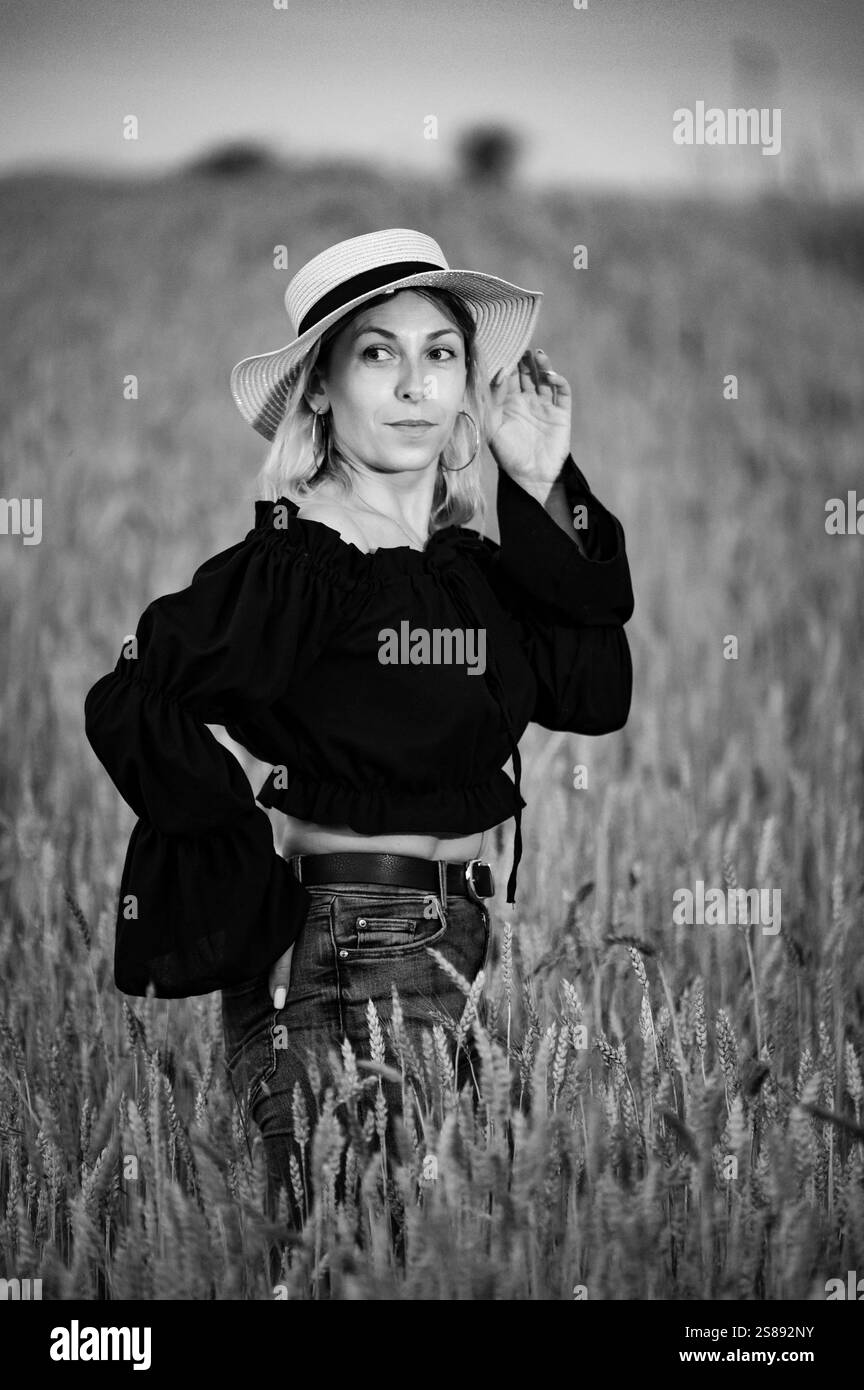 Ein schwarz-weißes Porträt einer Frau mit blondem Haar, mit Strohhut und schwarzem Oberteil, auf einem Feld aus hohem Weizen. Sie ist in einer ruhigen Mome gefangen Stockfoto