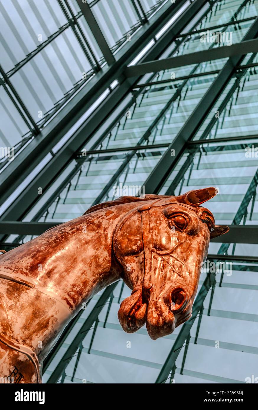 Eine auffällige Bronze-Pferdeskulptur steht vor einem modernen Glasatrium, das komplexe Design und urbane architektonische Kontraste zeigt. Stockfoto