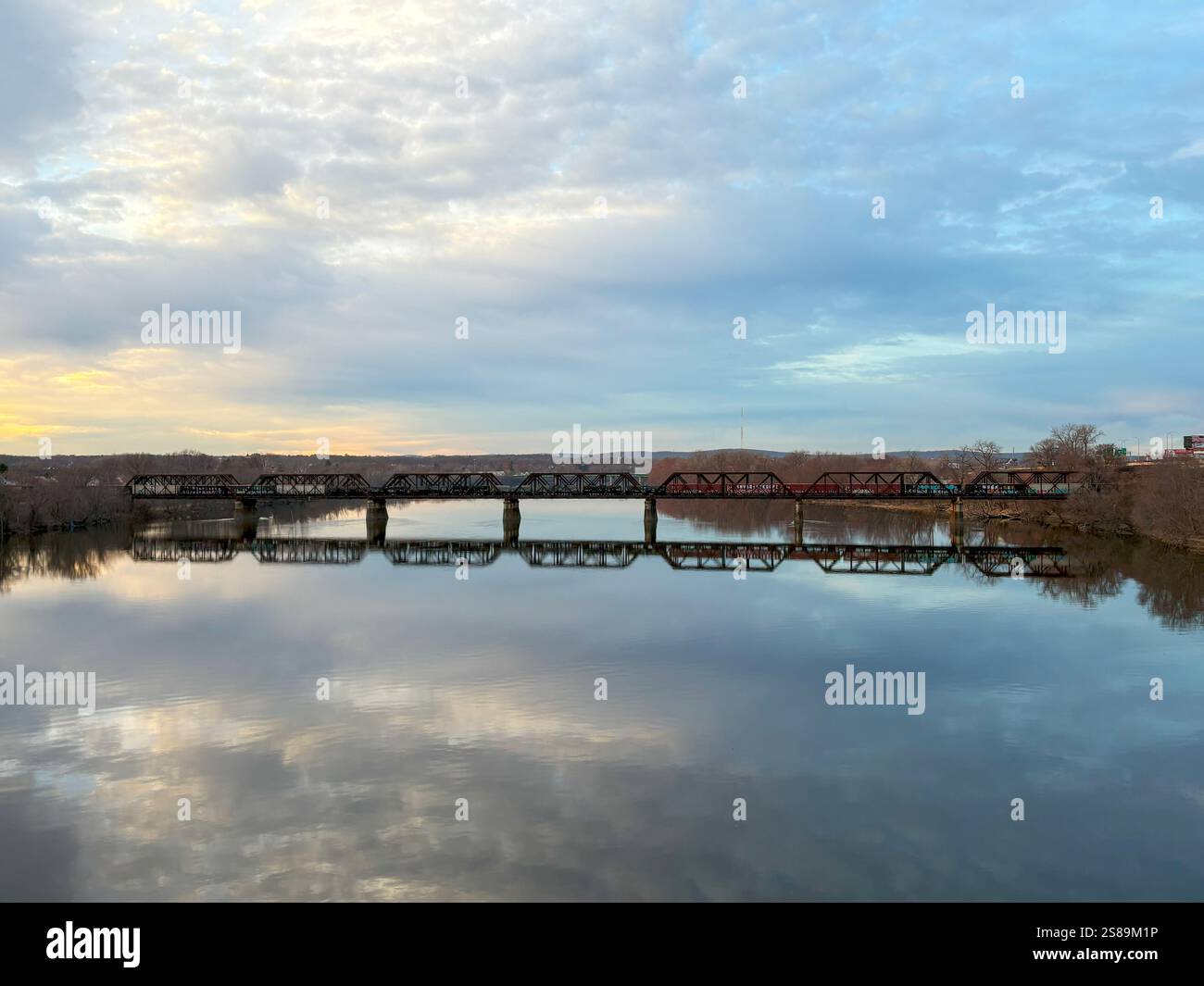 Eine Zugbrücke überspannt einen ruhigen Fluss mit sanften Farben des Sonnenuntergangs, die sich perfekt auf der Wasseroberfläche spiegeln. Stockfoto