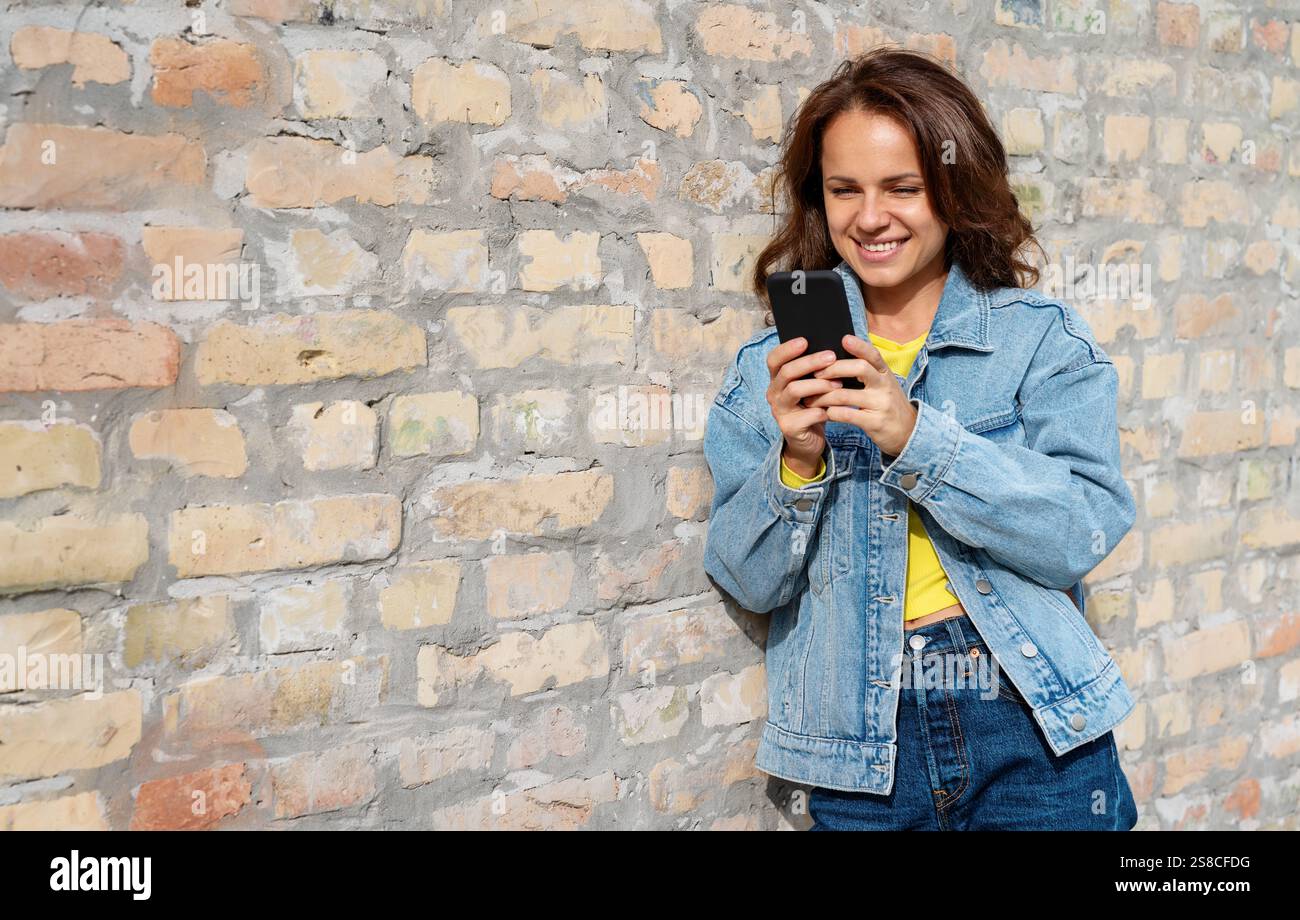 Junge städtische Frau in lässiger Kleidung steht draußen an einer Ziegelwand und schreibt auf ihrem Handy. Stockfoto