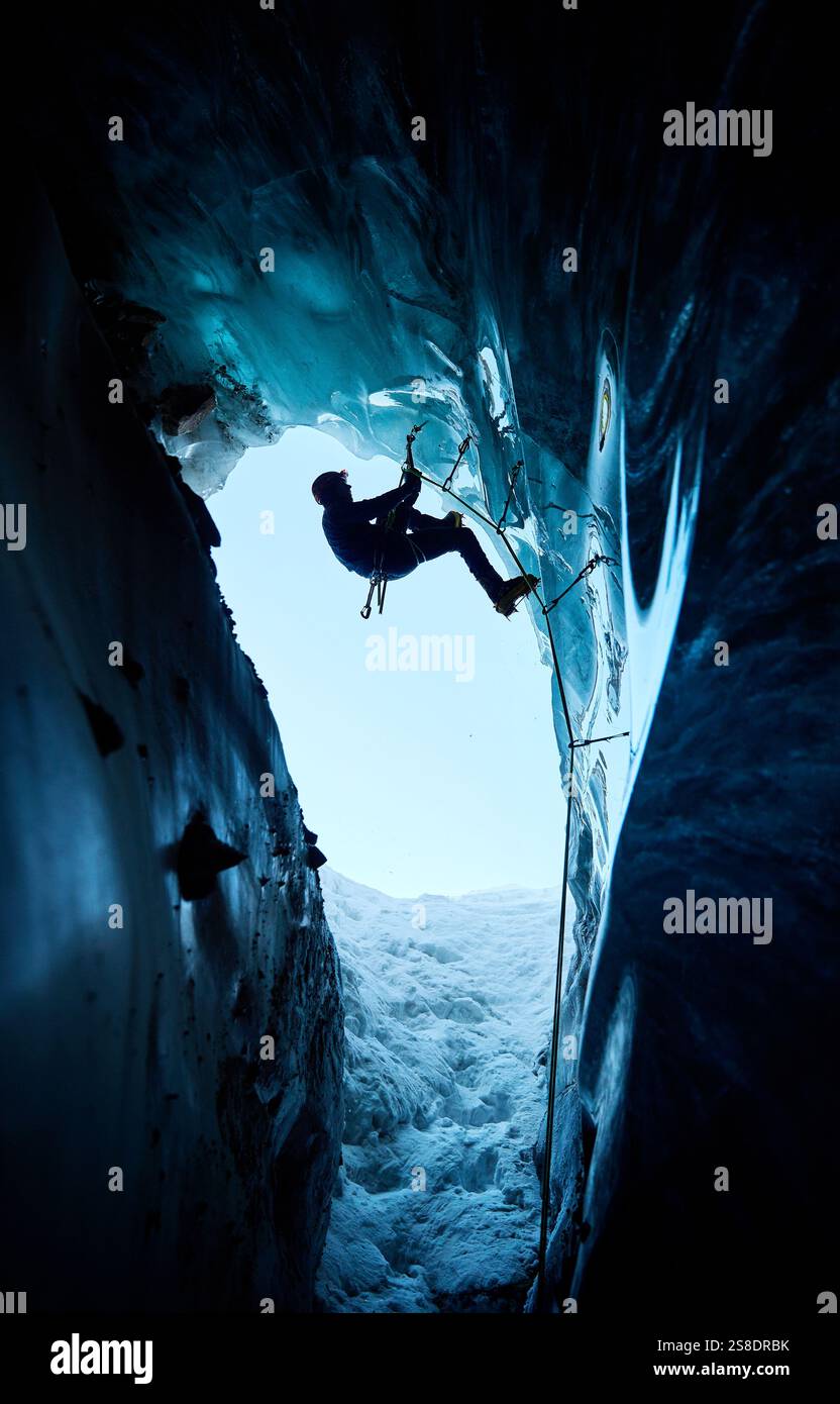Ein Mann in schwarzer Silhouette klettert auf eine Eishöhle im Inneren eines großen Gletschers mit Eisaxt und Steigeisen gegen blaue Ski in den Winterbergen. Sport-Mo Stockfoto