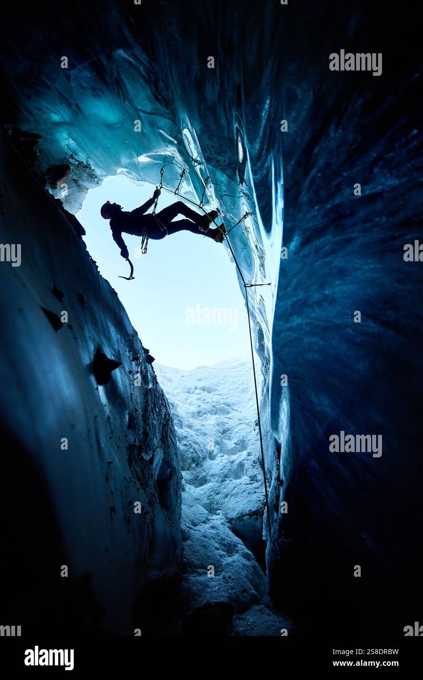 Ein Mann in schwarzer Silhouette klettert auf eine Eishöhle im Inneren eines großen Gletschers mit Eisaxt und Steigeisen gegen blaue Ski in den Winterbergen. Sport-Mo Stockfoto