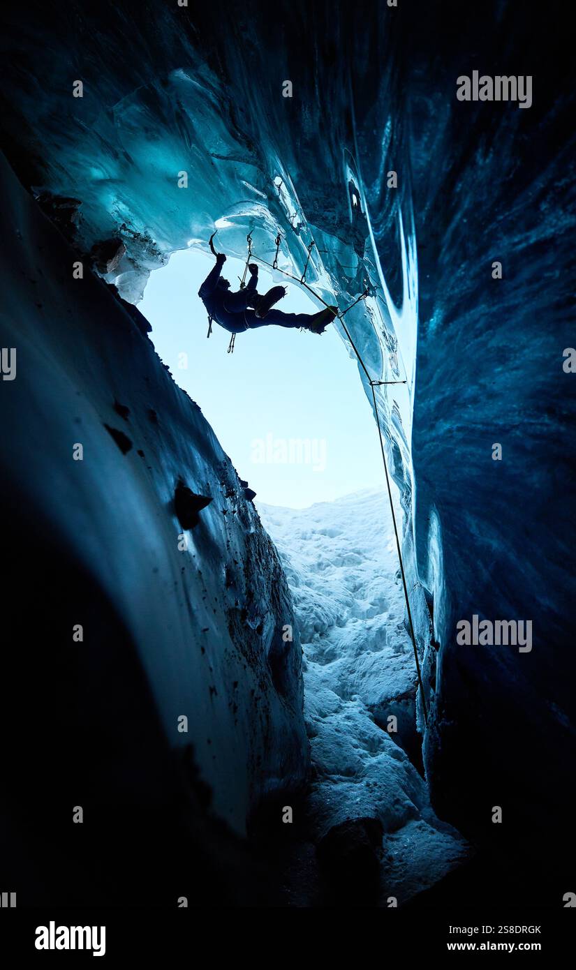 Ein Mann in schwarzer Silhouette klettert auf eine Eishöhle im Inneren eines großen Gletschers mit Eisaxt und Steigeisen gegen blaue Ski in den Winterbergen. Sport-Mo Stockfoto