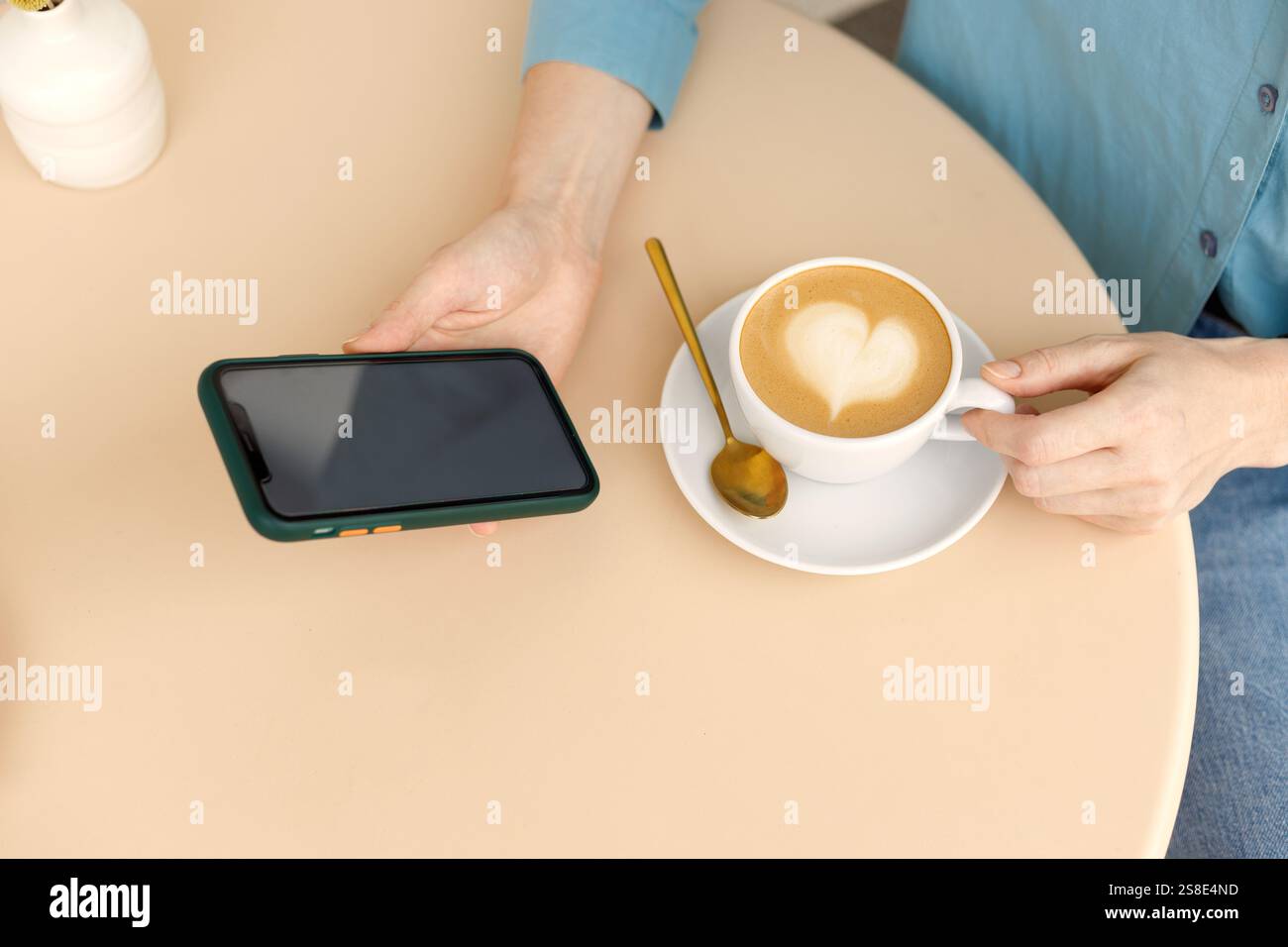 Eine Frau entspannt sich in einem Warschauer Café, hält ein Smartphone und eine Tasse Kaffee mit Latte Art in der Hand. Die gemütliche Umgebung und moderne Gadgets sorgen für eine friedliche Atmosphäre Stockfoto