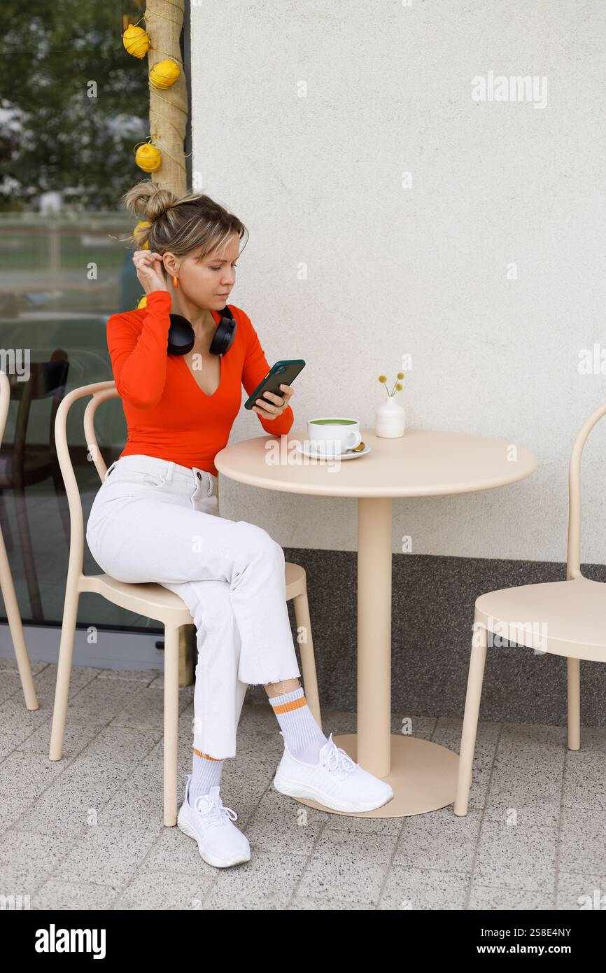 Junge Frau in leuchtend orangefarbenem Oberteil, sitzt in einem Café im Freien in Warschau Sie ist mit ihrem Smartphone und ihren Kopfhörern beschäftigt, während eine Tasse Kaffee an ist Stockfoto