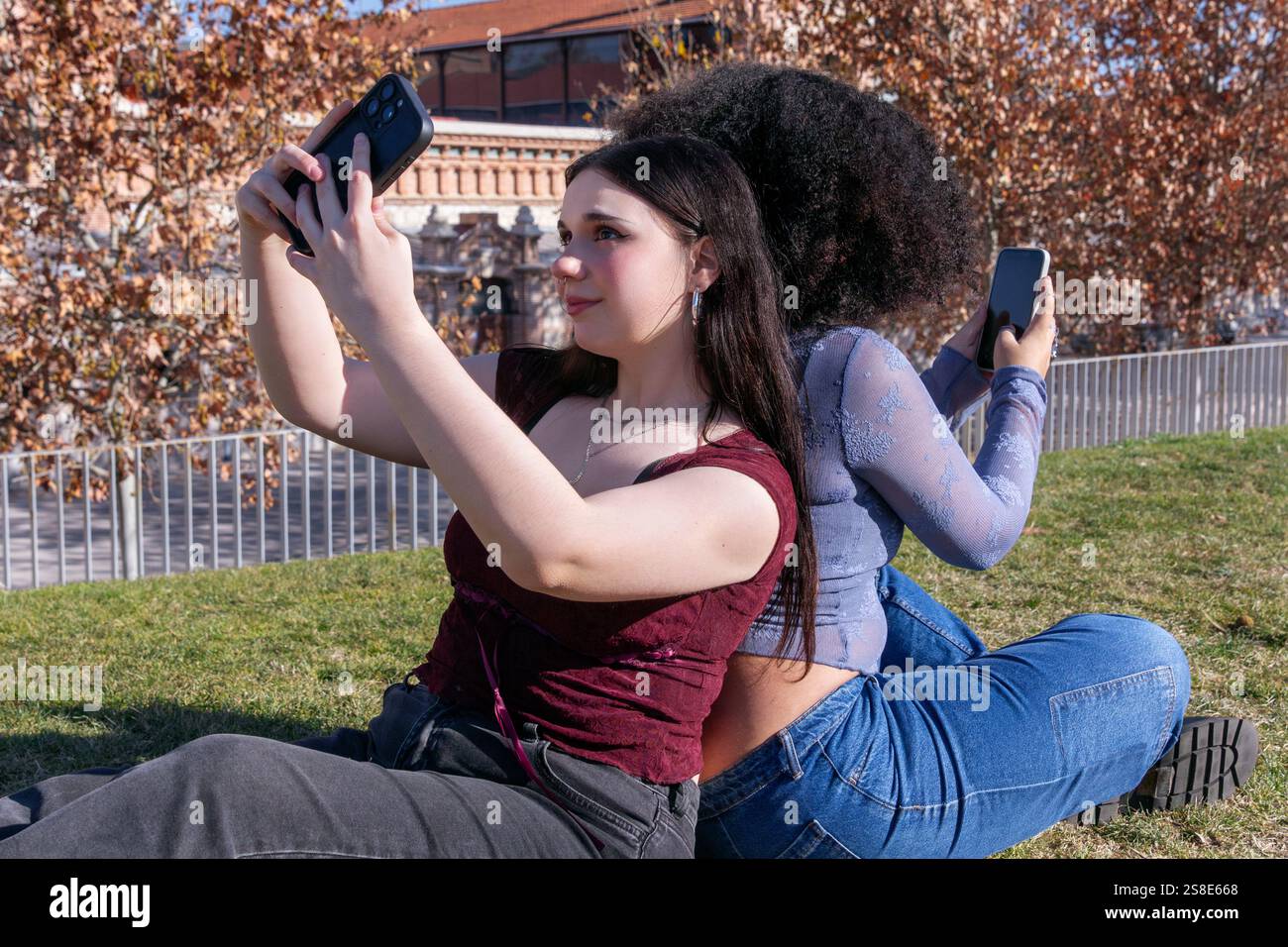 Zwei junge Frauen genießen einen sonnigen Tag im Freien, während sie Selfies machen. Die Dame links, mit hellem Haut, macht ein Foto mit ihrem Handy und ihrem Freund Stockfoto Zwei junge Frauen genießen einen sonnigen Tag im Freien, während sie Selfies machen. Die Dame links, mit hellem Haut, macht ein Foto mit ihrem Handy und ihrem Freund Stockfoto