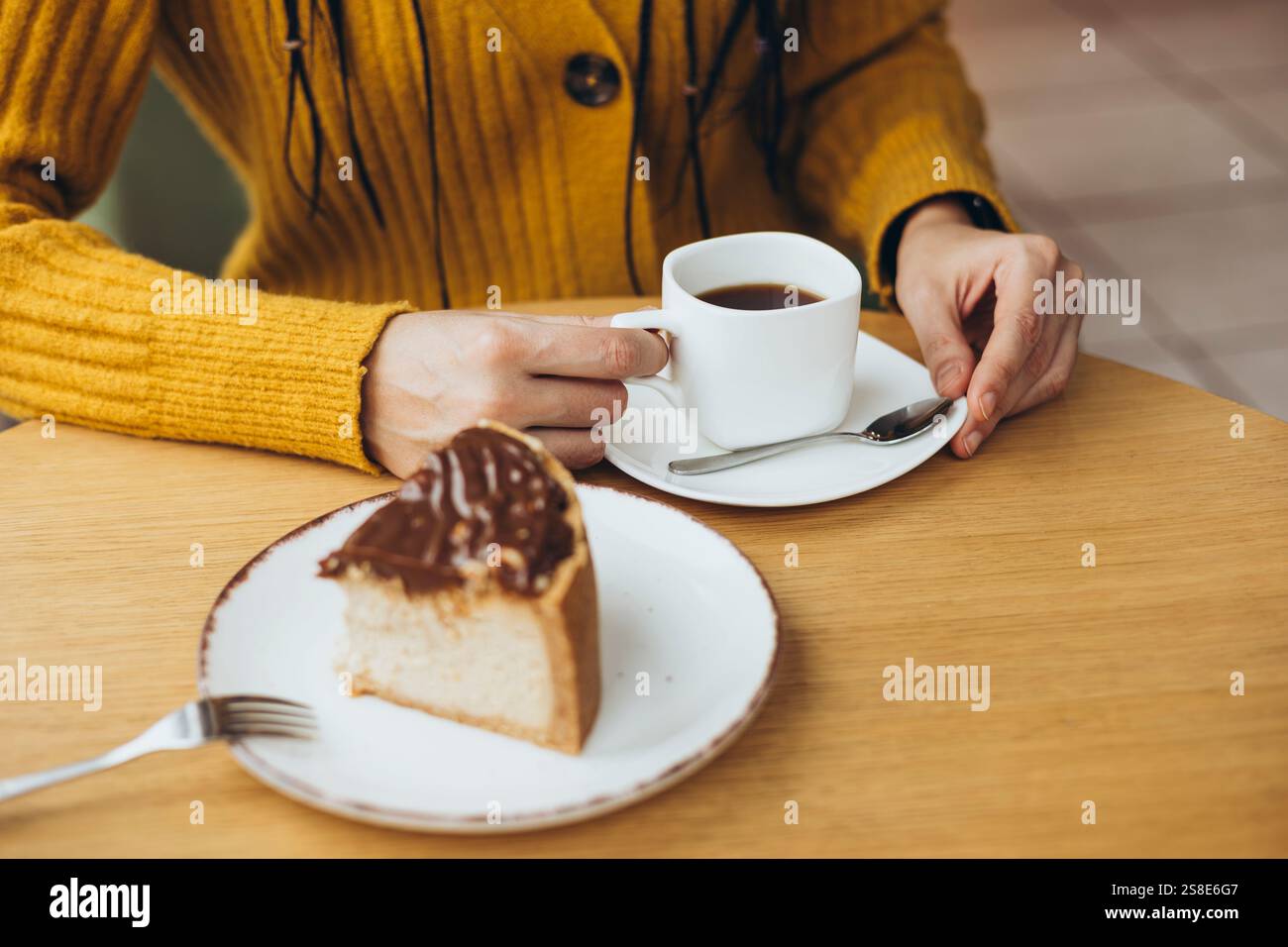 Eine Person in einem warmen gelben Pullover genießt eine ruhige Kaffeepause mit einer Tasse Kaffee und einem köstlichen Stück Schokoladenkuchen auf einem Holztisch Stockfoto