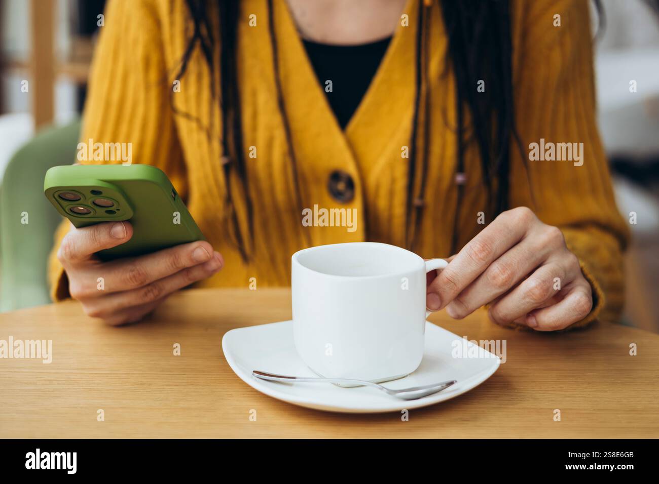 Eine Frau in einem gemütlichen gelben Pullover sitzt an einem Café-Tisch, hält eine weiße Kaffeetasse in der einen Hand und ein grünes Smartphone in der anderen und genießt eine entspannte Atmosphäre Stockfoto