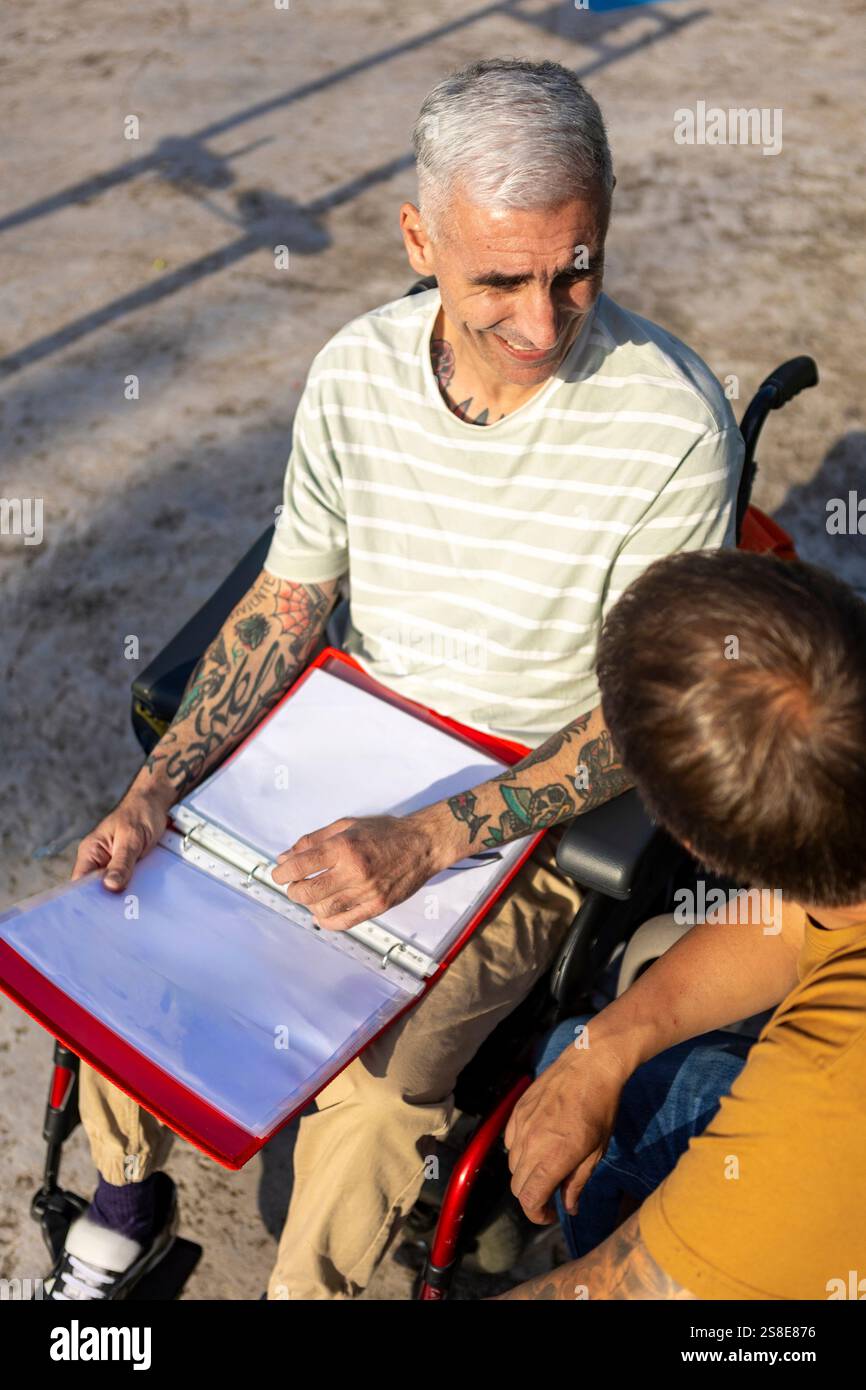 Zwei Männer führen ein freundliches Gespräch im Freien, einer sitzt im Rollstuhl mit Zerebralparese, hält einen offenen Ordner und zeigt einen animierten Ausdruck Stockfoto