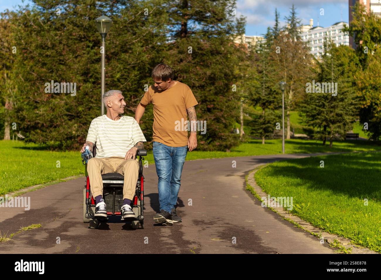Ein junger Mann läuft neben einem älteren Rollstuhlfahrer mit Zerebralparese in einem Park und führt zu freundlichen Gesprächen. Der Weg ist umgeben von Gree Stockfoto