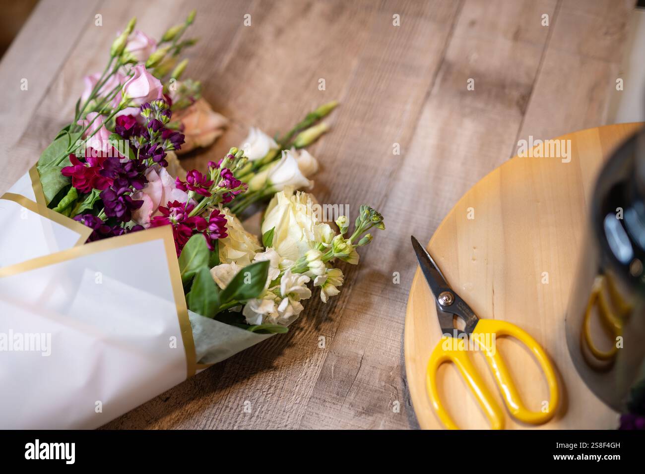 Schöne Auswahl an lebendigen Blumen, die in Papier gewickelt sind, mit einer gelben Schere in der Nähe auf einer Holzoberfläche, die Kreativität und Vorbereitung veranschaulicht Stockfoto