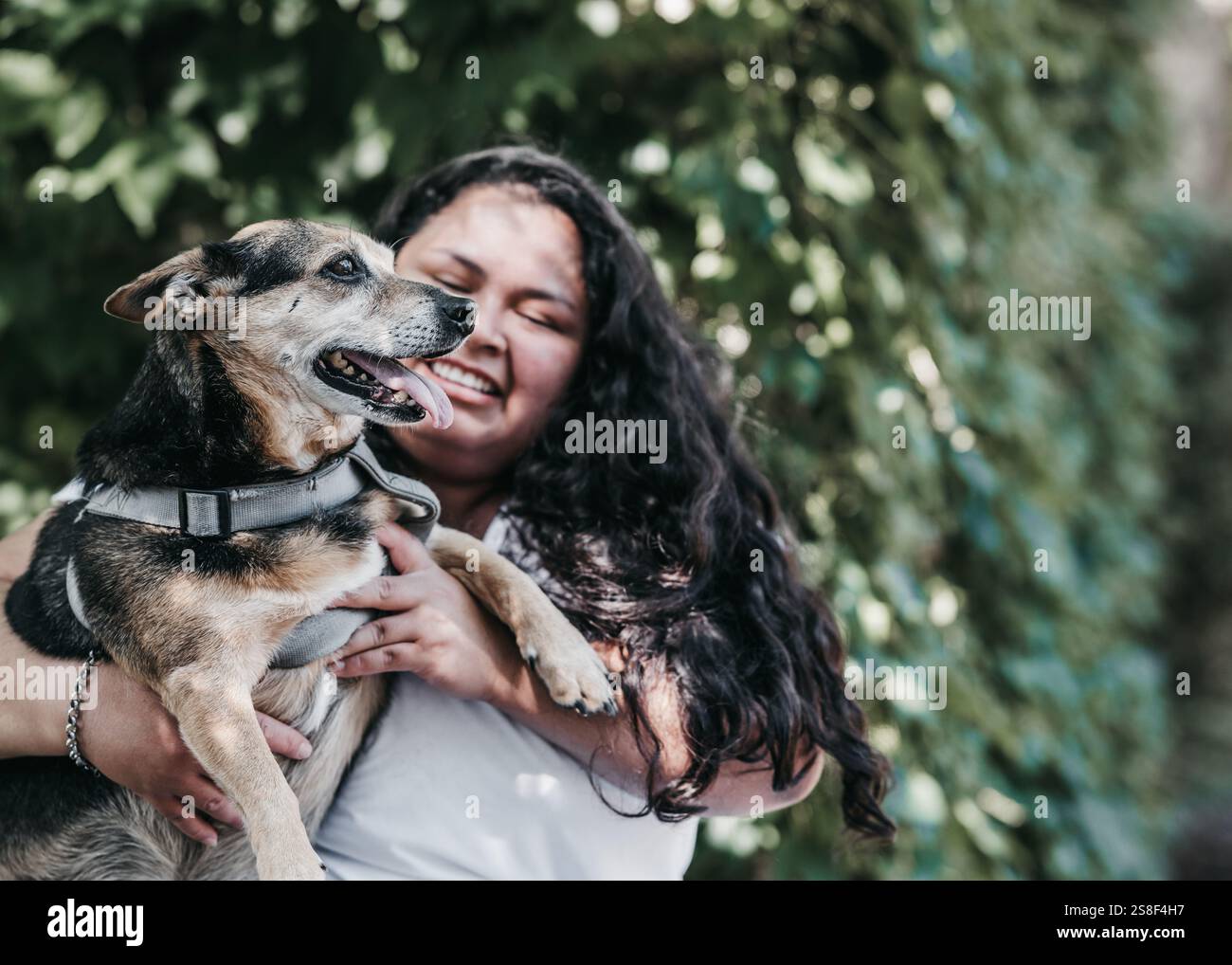 Latina-Mädchen hält einen mittelgroßen Hund in einem grauen Gurt. Sie hat lange dunkle Haare und trägt ein weißes T-Shirt. Hintergrund von grünem Laub im Freien Stockfoto