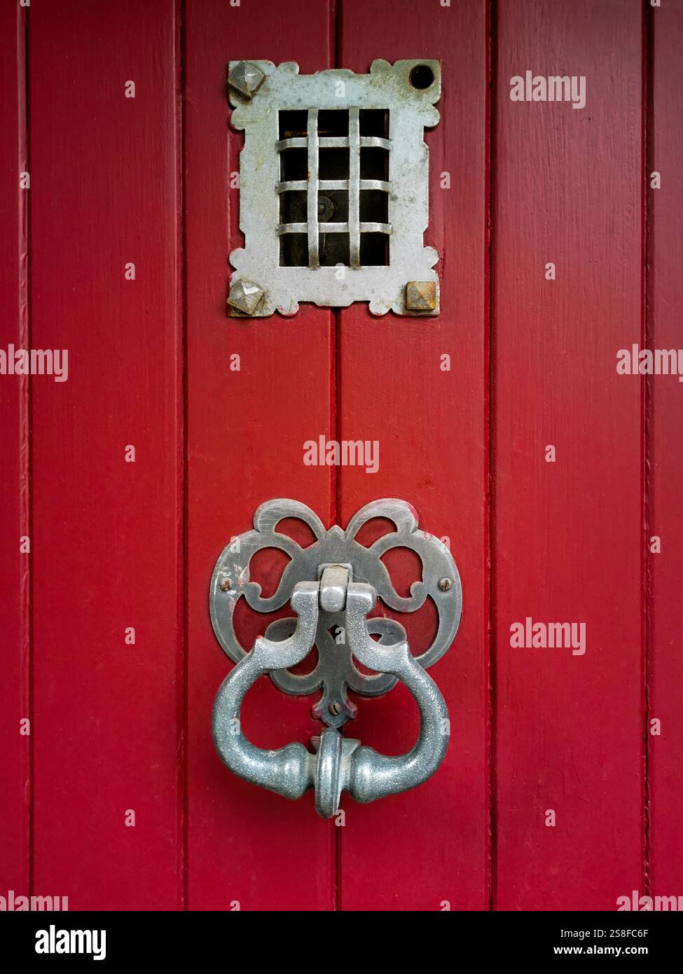 Eine auffällige rote Holztür zeigt komplizierte Metalldetails, darunter ein dekorativer Klopfer und ein kleines Gitterfenster oben. Stockfoto