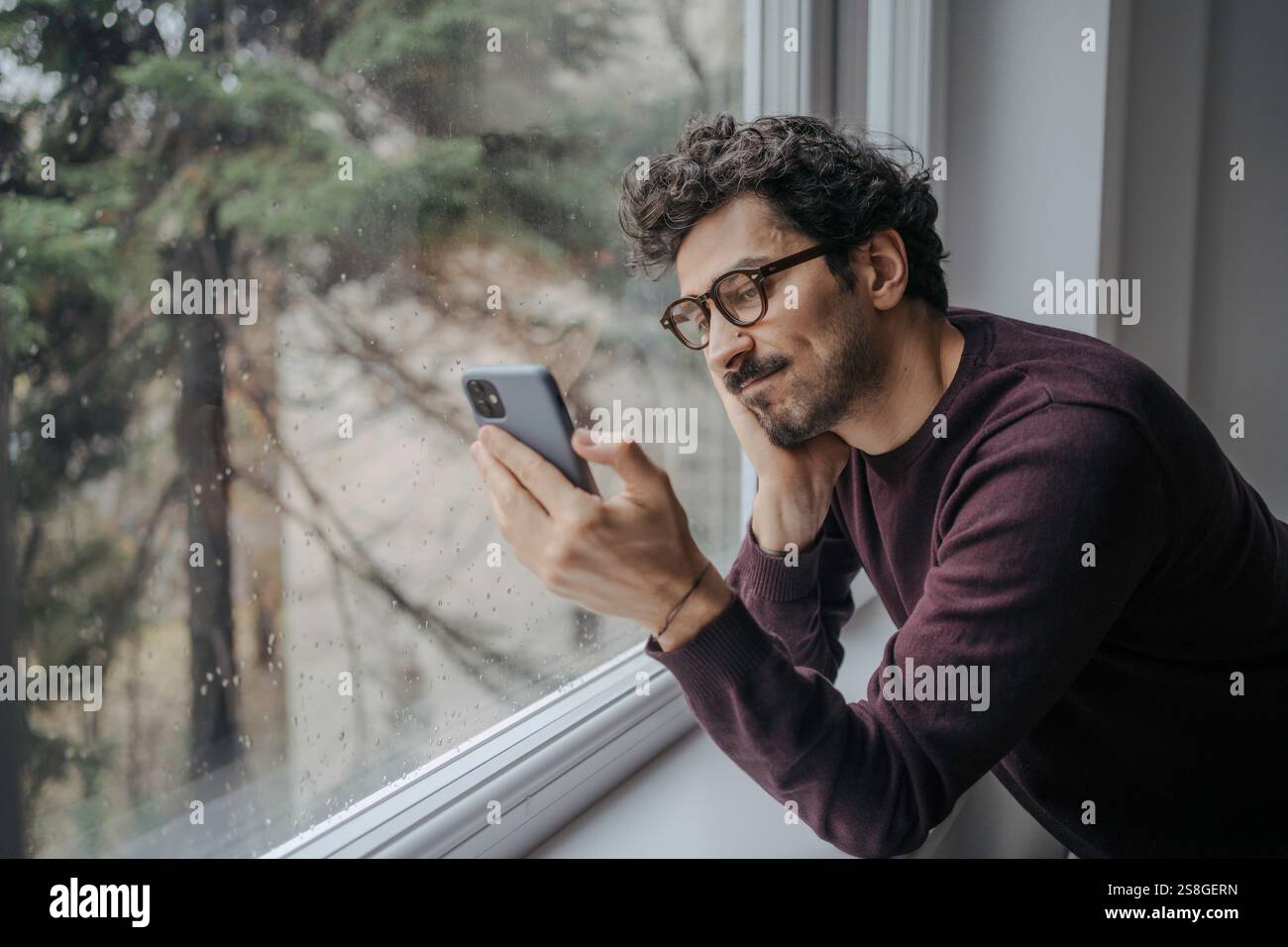 Junger gutaussehender Mann aus dem Nahen Osten mit Brille, der einen Videoanruf auf dem Handy hat, während er in der Nähe des Fensters steht. Stockfoto