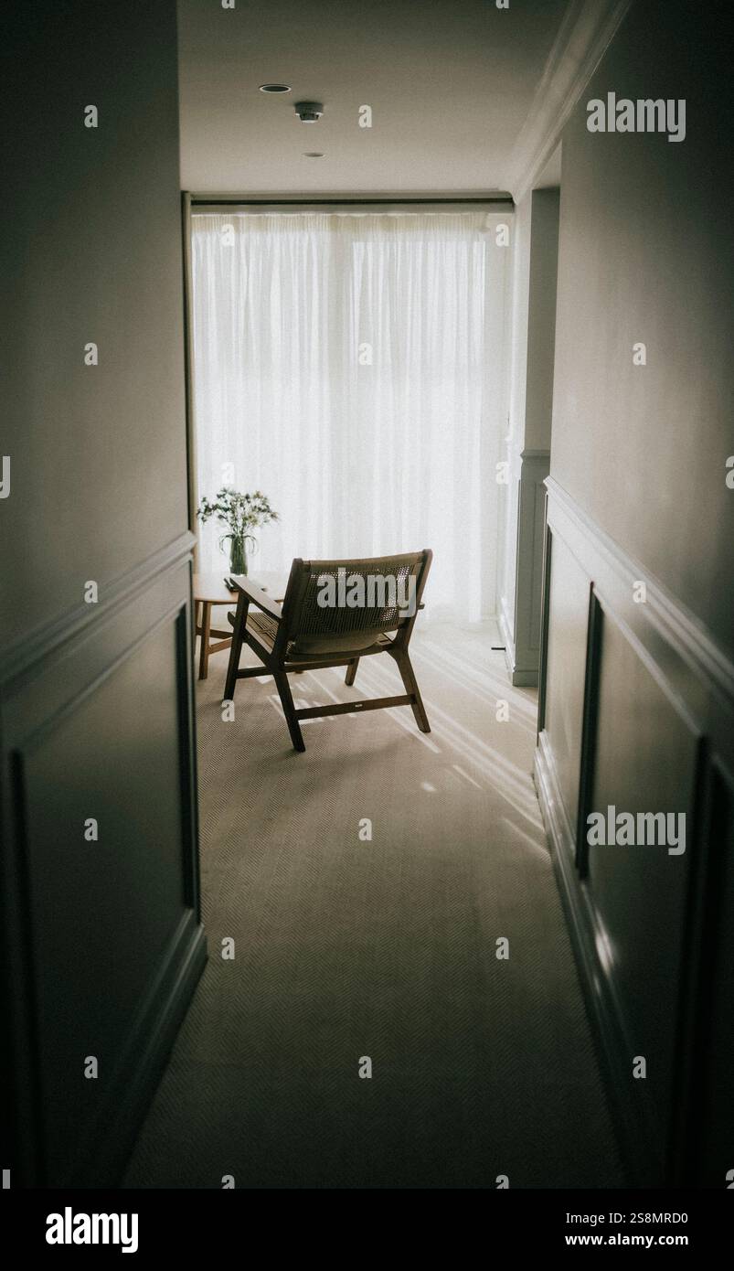 Ein Flur mit einem Stuhl und einem Tisch. Der Stuhl ist leer und auf dem Tisch ist eine Vase mit Blumen drauf. Das Zimmer ist schwach beleuchtet und die Atmosphäre ist ruhig Stockfoto