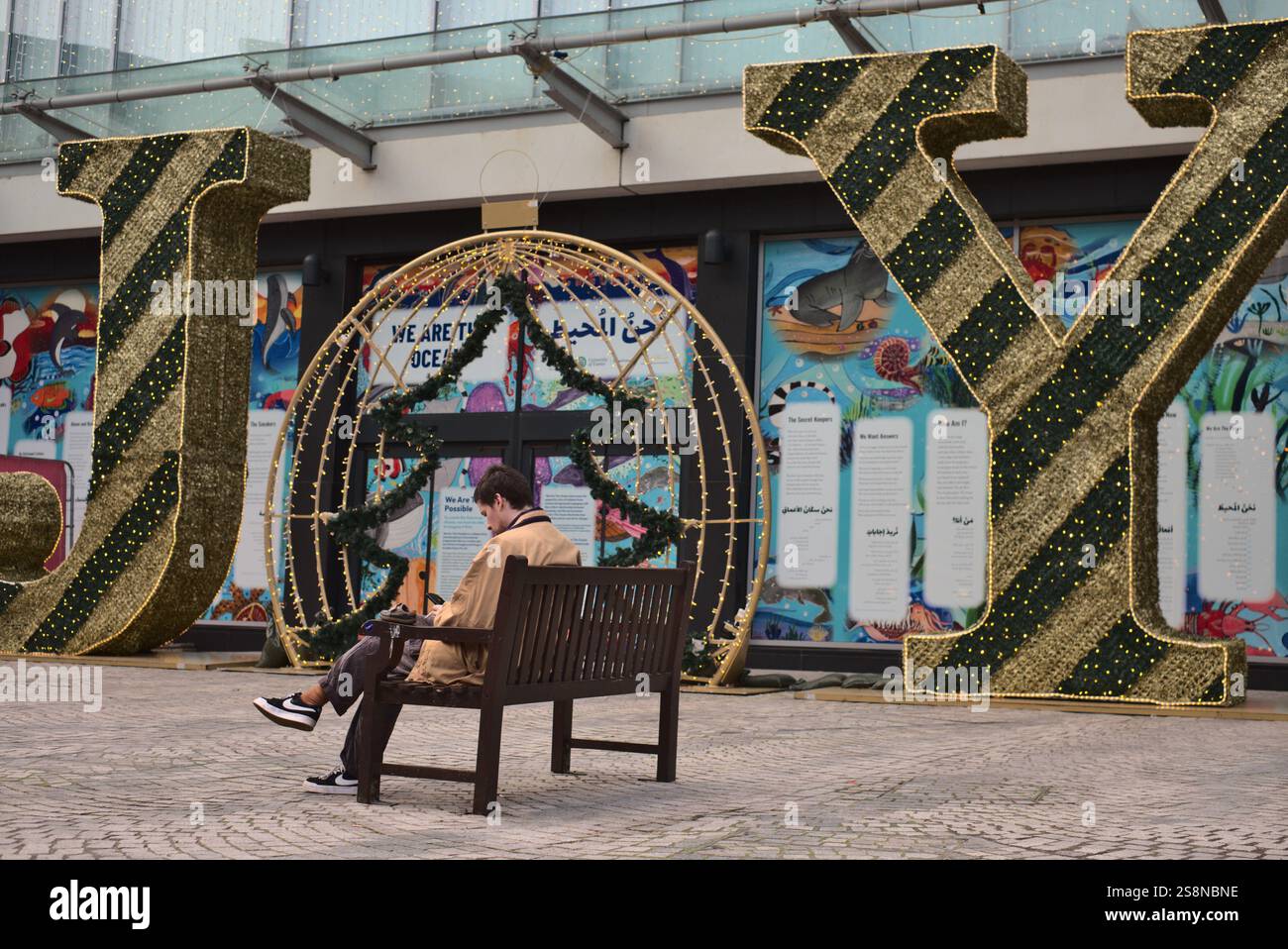 Ein junger Mann, der am Telefon surfte und eine Zigarette rauchte, saß auf einer öffentlichen Bank vor einer großen Weihnachtsdekoration mit dem Wort Joy. Exeter, Devon, Großbritannien Stockfoto