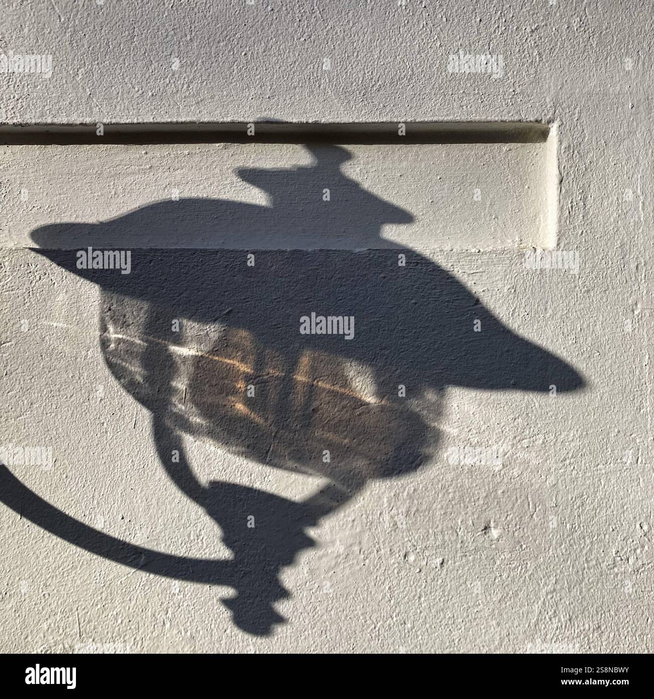 Schatten einer kunstvollen Vintage-Straßenlaterne, die unter Sonnenlicht scharf auf eine weiße strukturierte Wand gegossen wird. - Smartphone-aufgenommenes Stockfoto