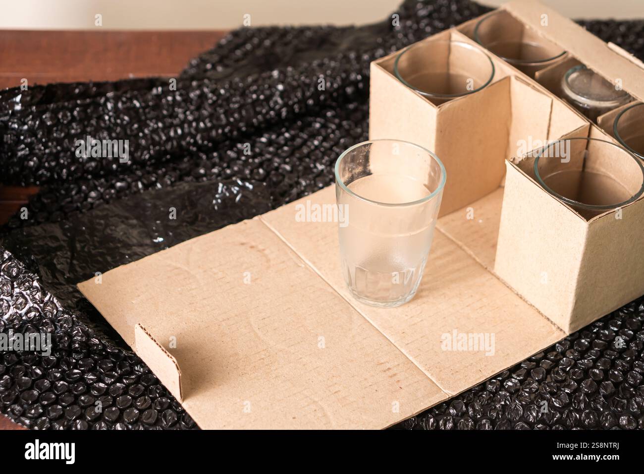 Ein klares Glas auf einer Pappoberfläche, umgeben von einer Schachtel mit weiteren Gläsern. Der Hintergrund ist mit schwarzer Luftpolsterfolie versehen, was auf Packag hindeutet Stockfoto