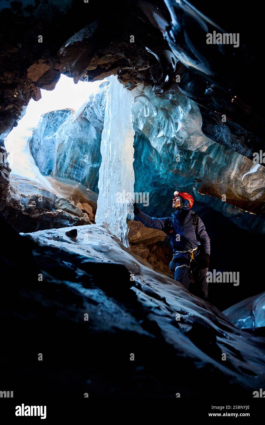 Man klettert in einer gefrorenen Eishöhle mit gefrorenem Wasserfall im Inneren des großen Gletschers mit Eisaxt und rotem Helm in den Winterbergen. Sportbergsteigen A Stockfoto