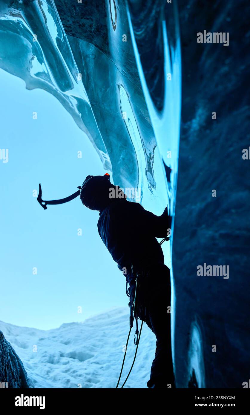 Ein Mann in schwarzer Silhouette klettert auf eine Eishöhle im Inneren eines großen Gletschers mit Eisaxt und Steigeisen gegen blaue Ski in den Winterbergen. Sport-Mo Stockfoto