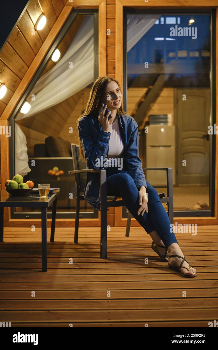 Niedliche junge Frau, die am späten Abend auf der Terrasse eines Bungalows sitzt und mit dem Smartphone spricht Stockfoto