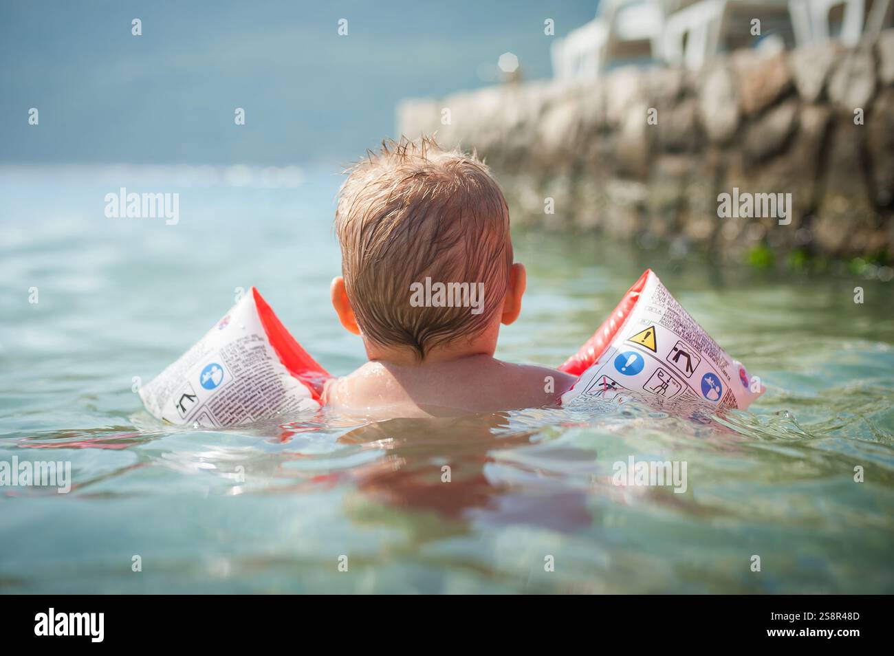 Rückansicht eines Kleinkindes lernt gern in klarem, seichten Wasser schwimmen, während es Schwimmhilfen trägt. Die Sonne scheint hell, während sie ihre erkunden Stockfoto