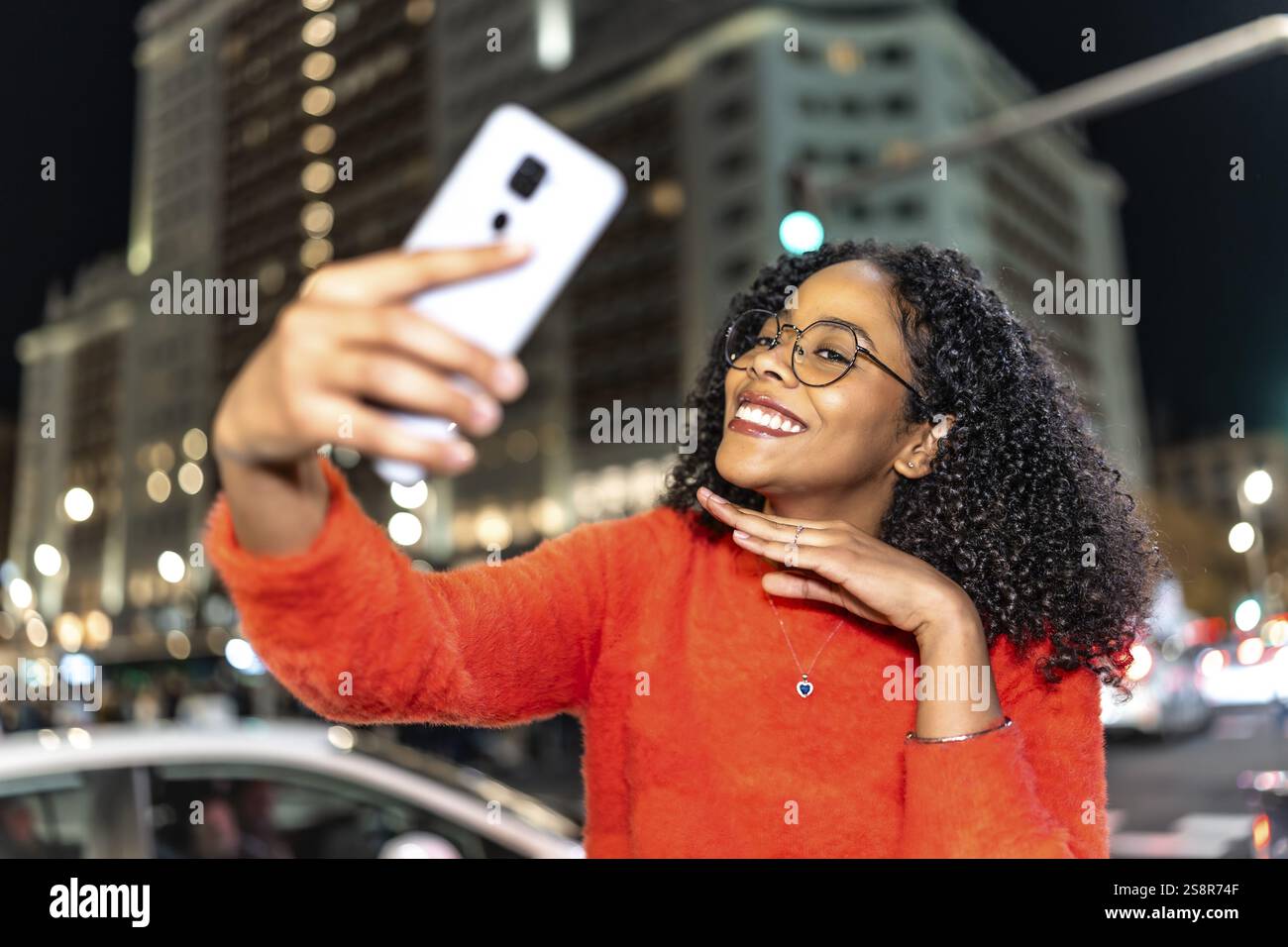 Fröhliche junge Frau lächelt, während sie mit ihrem Smartphone ein Selfie in einer lebhaften Stadt bei Nacht, umgeben von wunderschönen Lichtern, macht Stockfoto