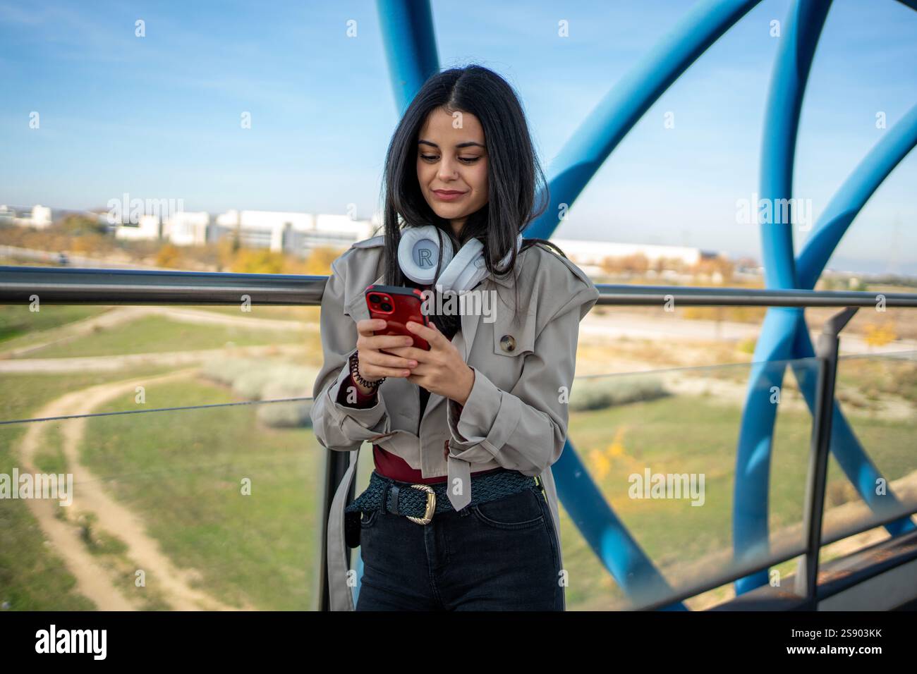 Stilvolle junge Frau, die auf einer modernen Brücke mit einem Smartphone steht und die Aussicht auf die Stadt und die Konnektivität genießt Stockfoto