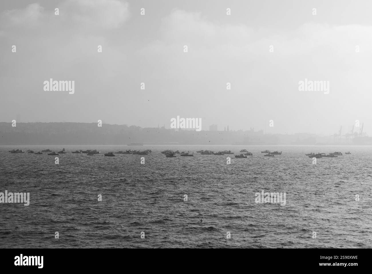Ein schwarz-weißer atmosphärischer Blick auf Fischerboote, die auf dem Meer schwimmen, mit einer trüben Skyline der Stadt im Hintergrund Stockfoto
