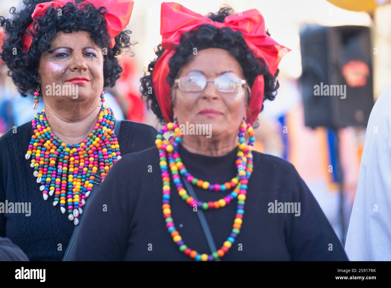 Zwei Personen mit festlicher Kleidung, farbenfrohen Perlen und roten Haarschleifen bei einer lebhaften Veranstaltung. Einer rechts im Fokus, mit Brille und einer ernsthaften Stockfoto