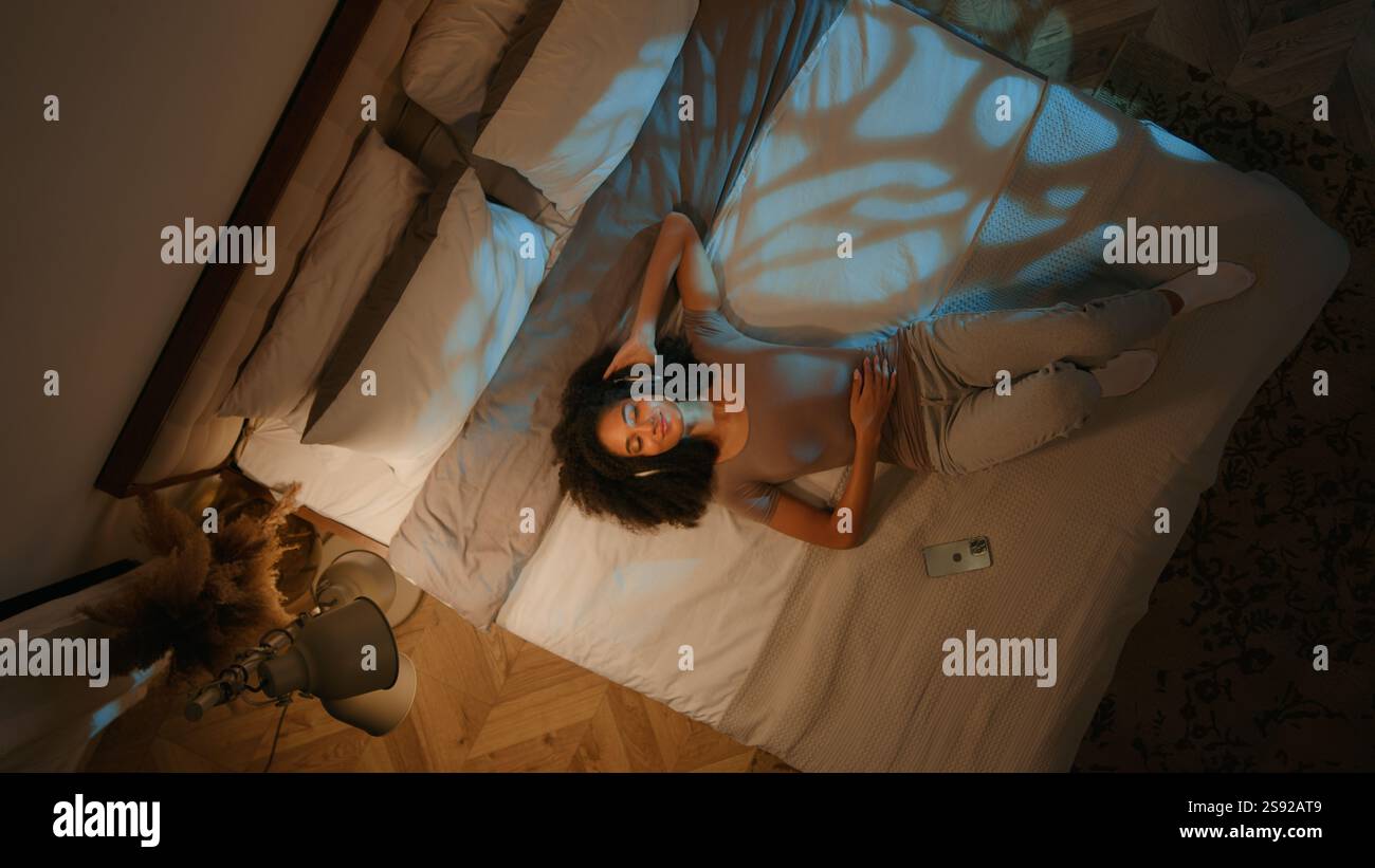Blick von oben entspanntes Mädchen, das auf ein komfortables Bett fällt Nacht Freizeit Musik im Kopfhörer hört Liedaugen geschlossen entspannter Abend Stockfoto