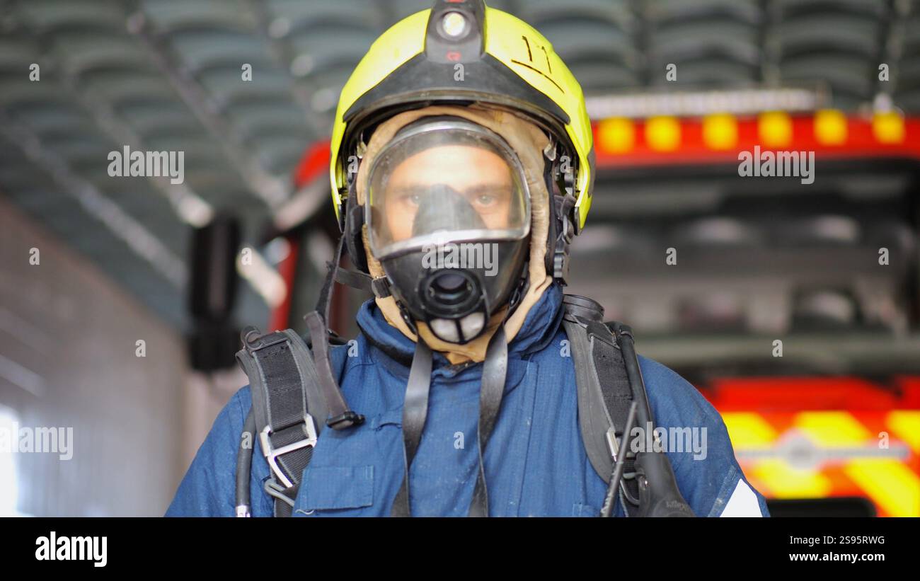 Porträt eines männlichen Feuerwehrmannes in Schutzhelm und Uniform, der in der Nähe eines großen roten Autos auf der Station steht. Junger selbstbewusster Feuerwehrmann in voller Ausrüstung posiert Stockfoto