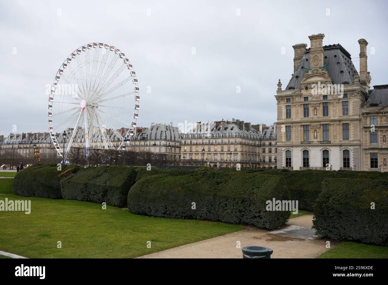 Ein malerischer Blick auf den Tuileriengarten mit Riesenrad, gepflegten Hecken, historischer Architektur und dem Louvre unter bewölktem Himmel. Stockfoto