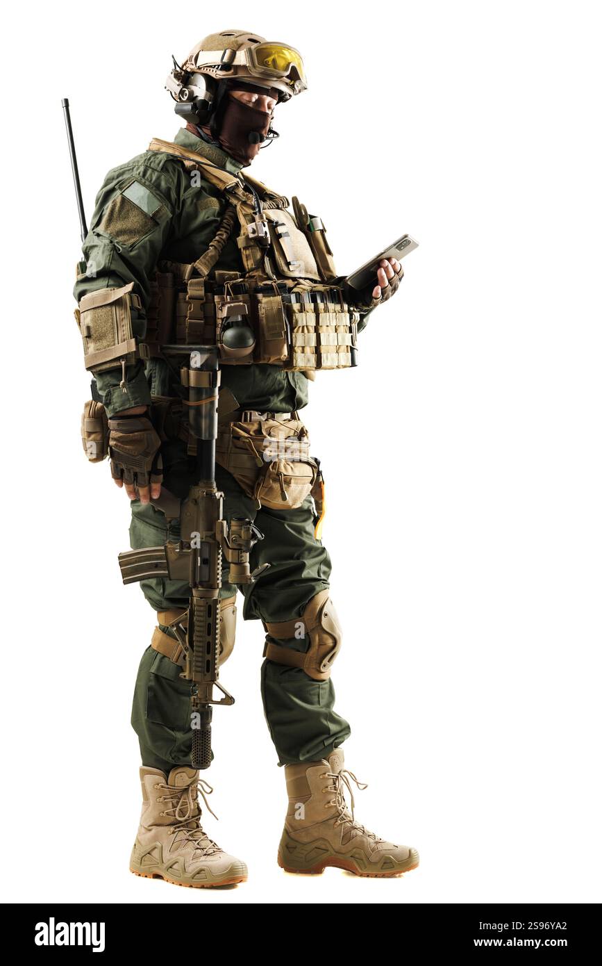 Ein bewaffneter Soldat in voller taktischer Ausrüstung, der ein Handgerät untersucht und fortschrittliche militärische Ausrüstung und Vorsorge zeigt. Das Bild wird hervorgehoben Stockfoto