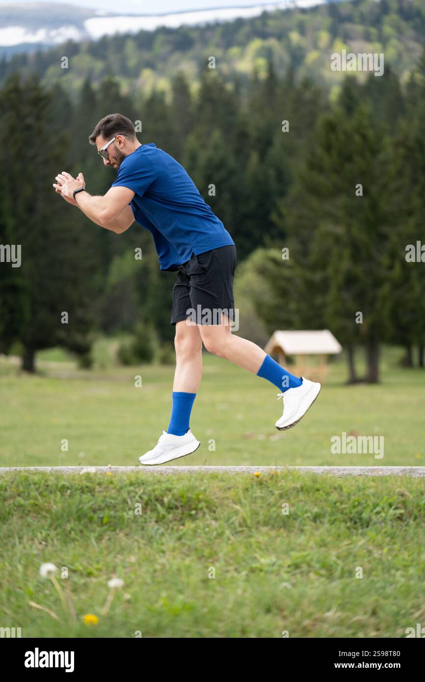 Mann im sportlichen Outfit steht fokussiert und bereit für Einen Sprint, eingerahmt von Green Grass, Bäumen und Einem Bergblick Stockfoto