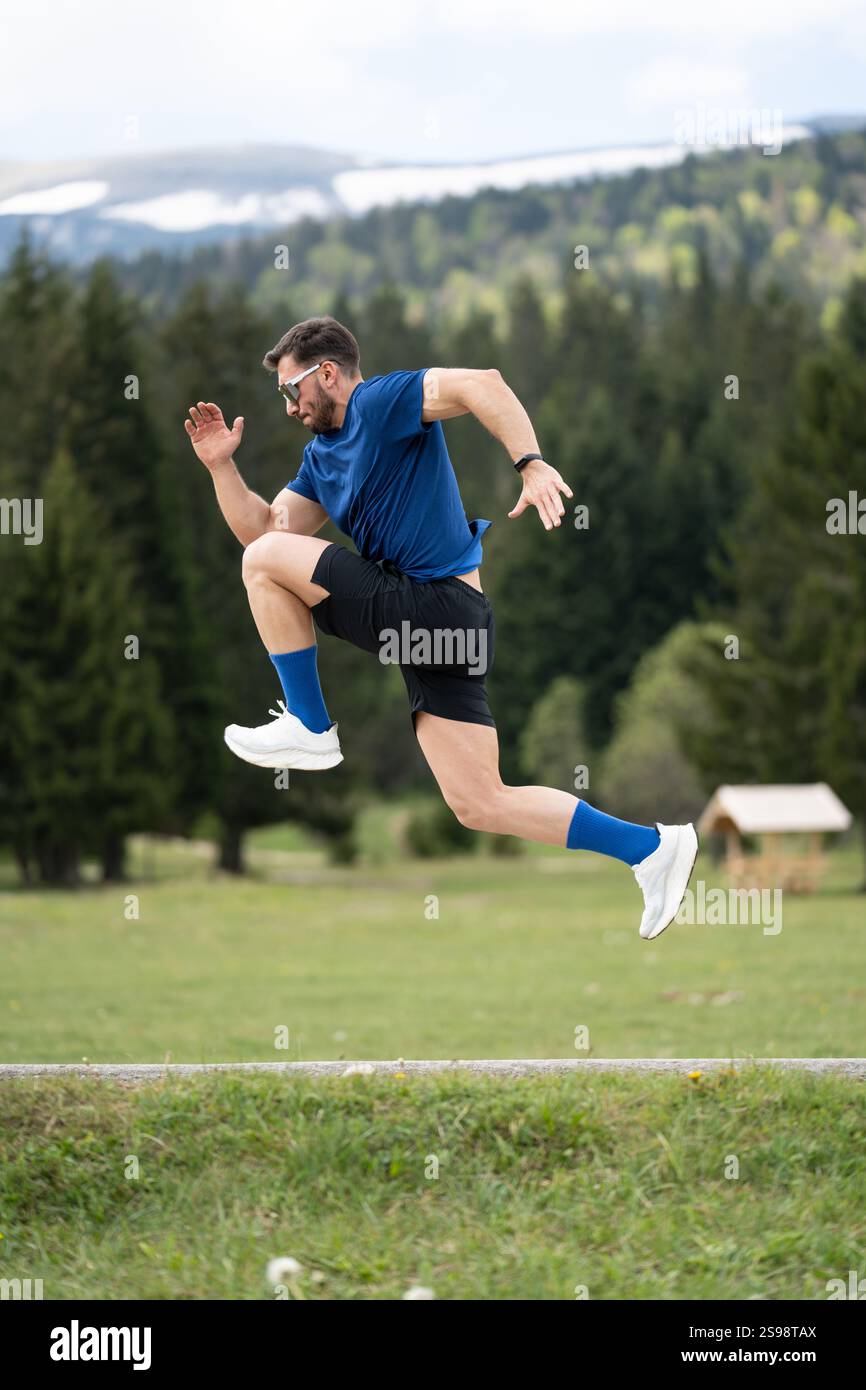 Mann im sportlichen Outfit steht fokussiert und bereit für Einen Sprint, eingerahmt von Green Grass, Bäumen und Einem Bergblick Stockfoto