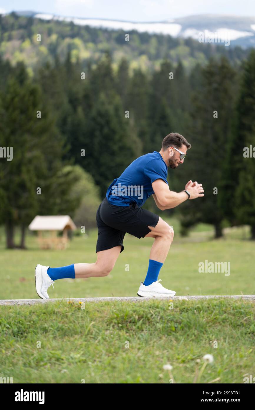 Mann im sportlichen Outfit steht fokussiert und bereit für Einen Sprint, eingerahmt von Green Grass, Bäumen und Einem Bergblick Stockfoto