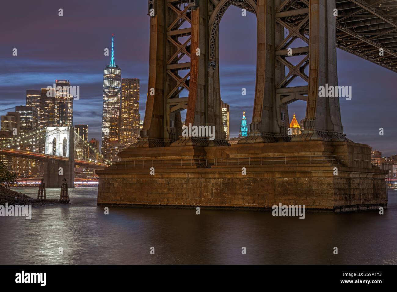 Einer der Brückenpfeiler der Manhattan Bridge in New York bei Nacht mit der Brooklyn Bridge und Manhattan hinten Stockfoto