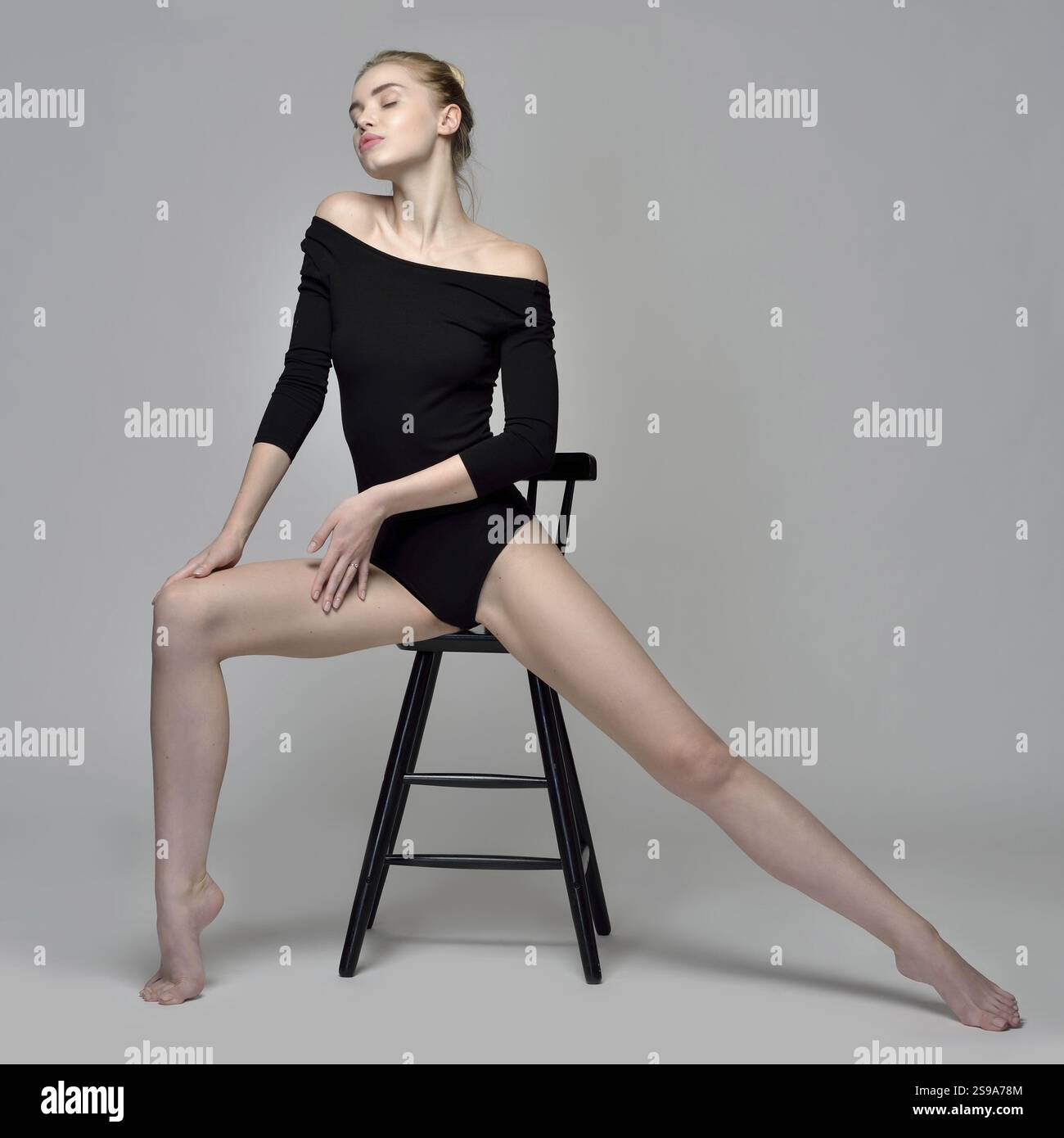 Schöne blonde Turnerin in schwarzem Kostüm. Sie sitzt auf einem Hochstuhl im Studio mit grauem Hintergrund, St. Petersburg, Russische Föderation Stockfoto