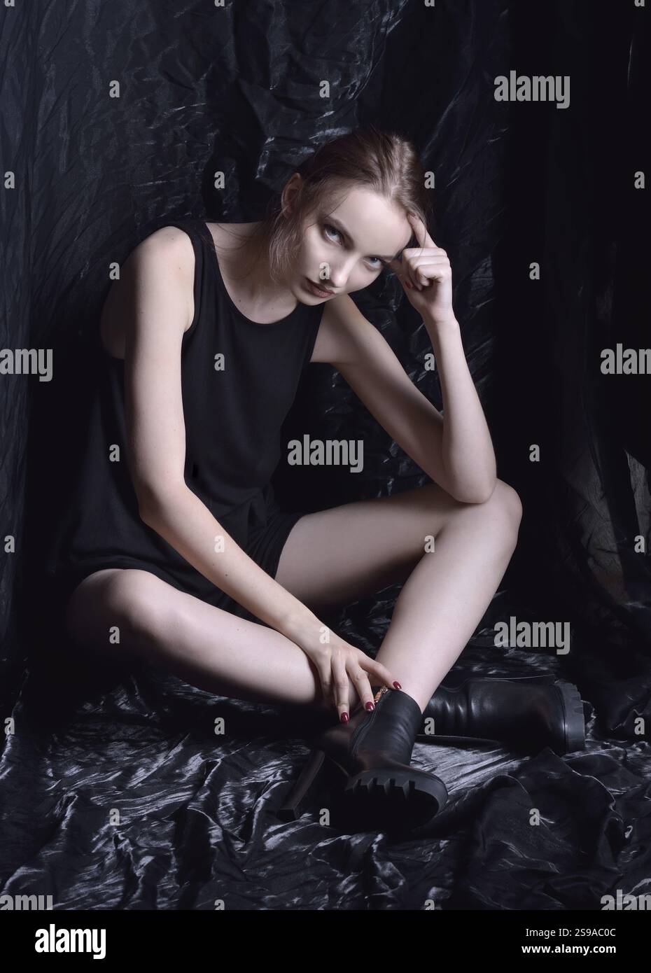 Sitzende schöne Frau in schwarzem Oberteil und Schuhen im Studio mit dunklem Hintergrund, St. Petersburg, Russische Föderation Stockfoto