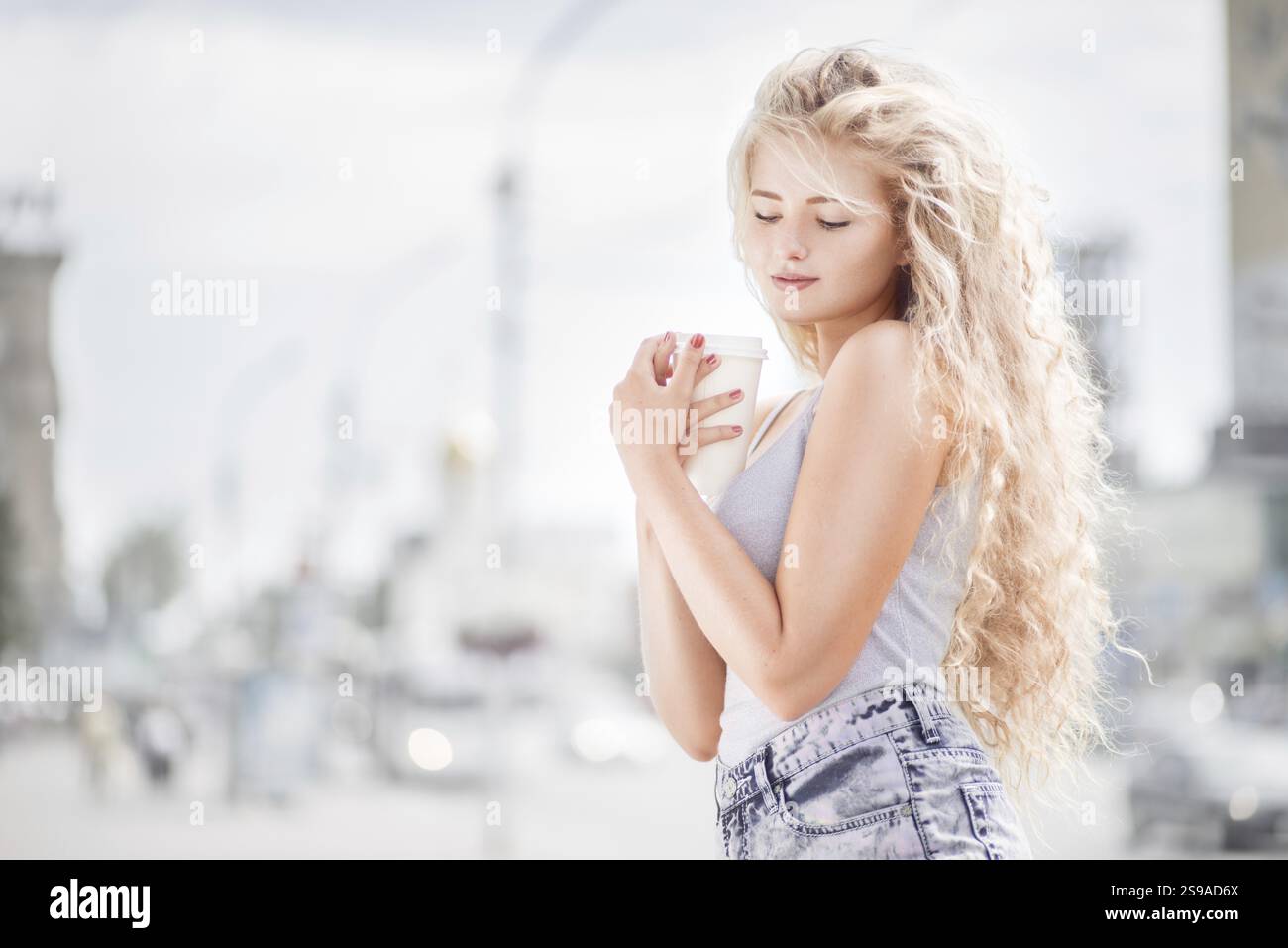Glückliche junge Frau mit langen lockigen Haaren, die eine Kaffeetasse zum Mitnehmen hält, lächelt und flirtet mit der Kamera vor dem Hintergrund des Stadtverkehrs Stockfoto Glückliche junge Frau mit langen lockigen Haaren, die eine Kaffeetasse zum Mitnehmen hält, lächelt und flirtet mit der Kamera vor dem Hintergrund des Stadtverkehrs Stockfoto