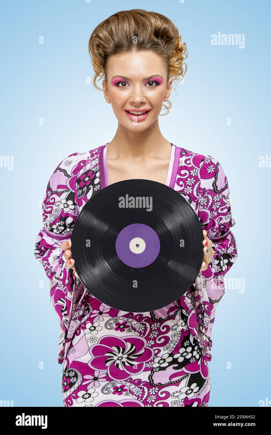 Farbenfrohes Foto einer fröhlichen, modischen Hippie-Hausfrau mit einer Retro-Schallplatte in den Händen auf blauem Hintergrund Stockfoto