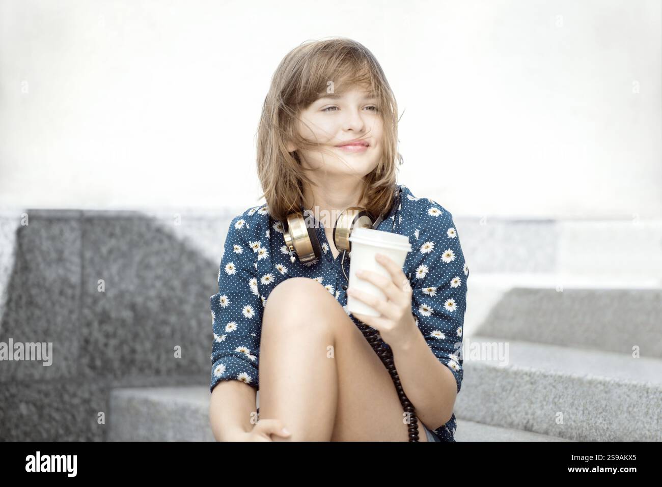 Glückliche junge Frau mit Vintage-Kopfhörern um den Hals, lächelt glücklich und sitzt auf einer Treppe mit einer Kaffeetasse zum Mitnehmen, vor einem städtischen bac Stockfoto