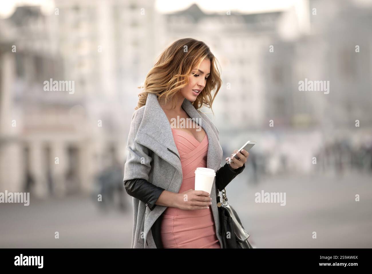 Eine Geschäftsfrau, die E-Mails auf ihrem Handy liest und eine Kaffeetasse in der Hand hält, vor einem städtischen Hintergrund Stockfoto