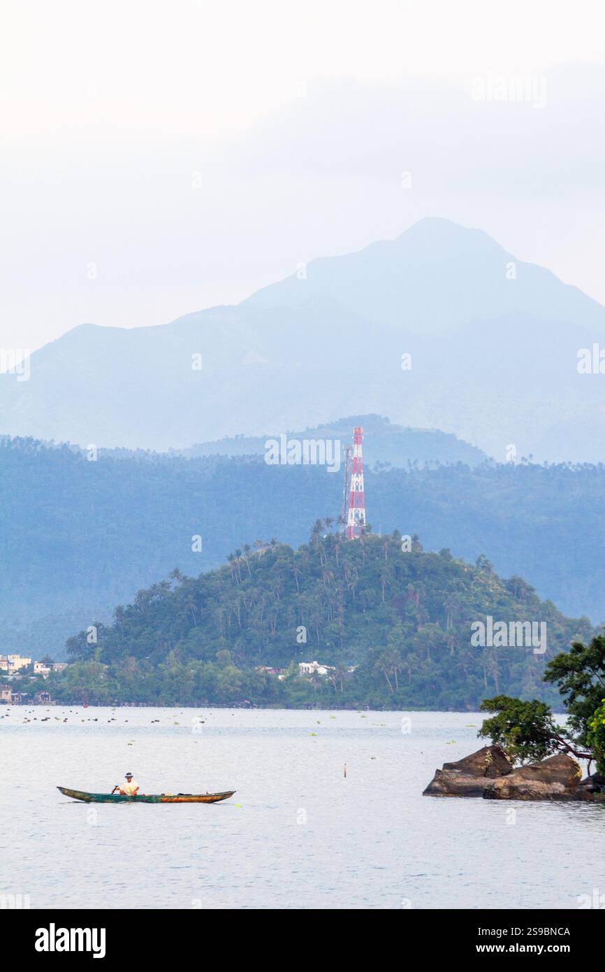 Malerischer Blick auf den Buhi-See in Bicol, Philippinen, mit einem Mann, der ein kleines Boot im Vordergrund paddelt, und üppige Berge im Hintergrund Stockfoto
