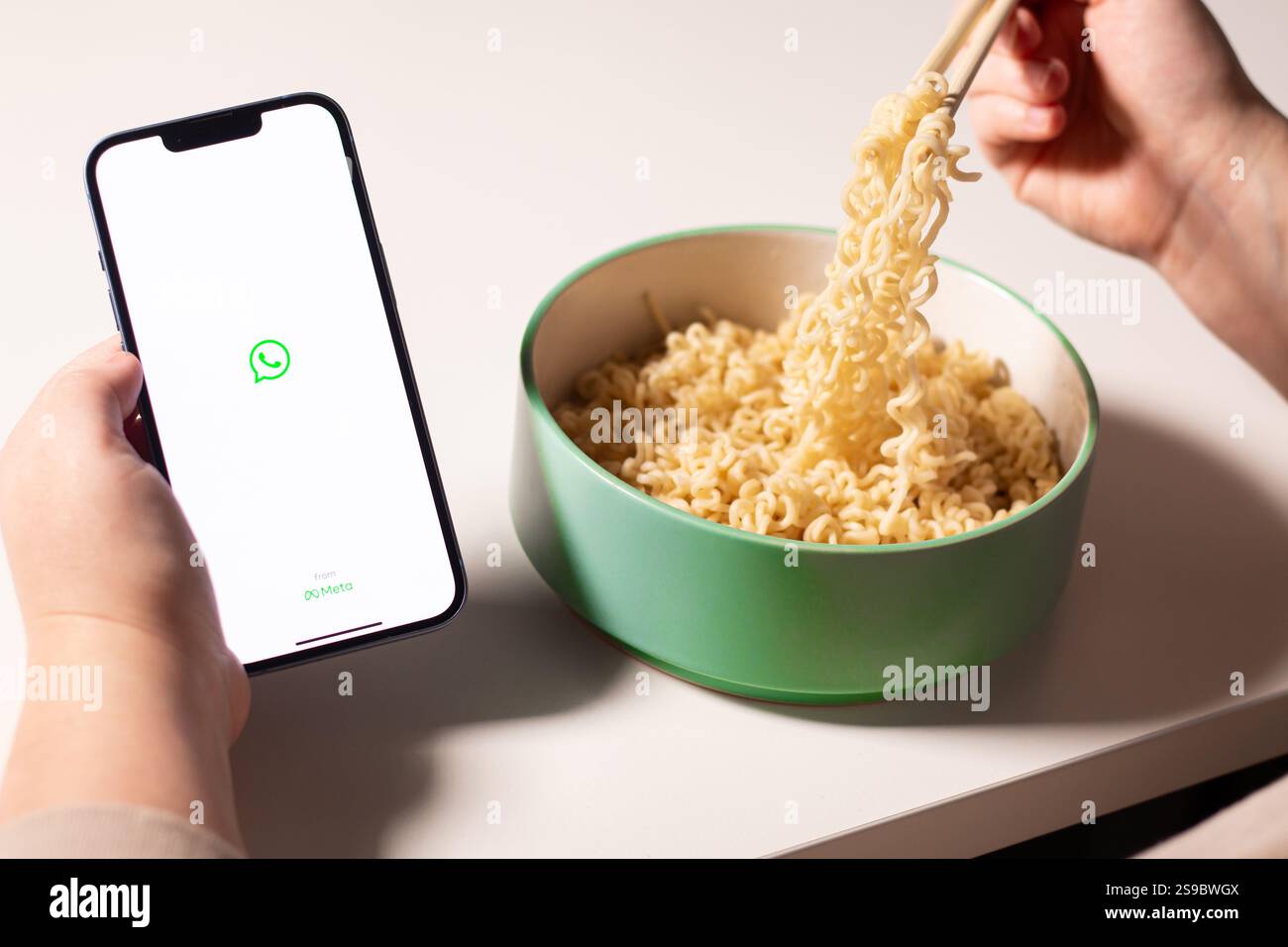 Frau, die die Whatsapp App auf dem Smartphone nutzt, während sie zu Hause Nudeln isst. WhatsApp-Logo auf dem Bildschirm. Essen mit Social-Media-Konzept Stockfoto